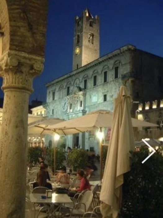 Outdoor seating area with tables, chairs, and umbrellas in front of a historic stone building with a clock tower, illuminated at night.