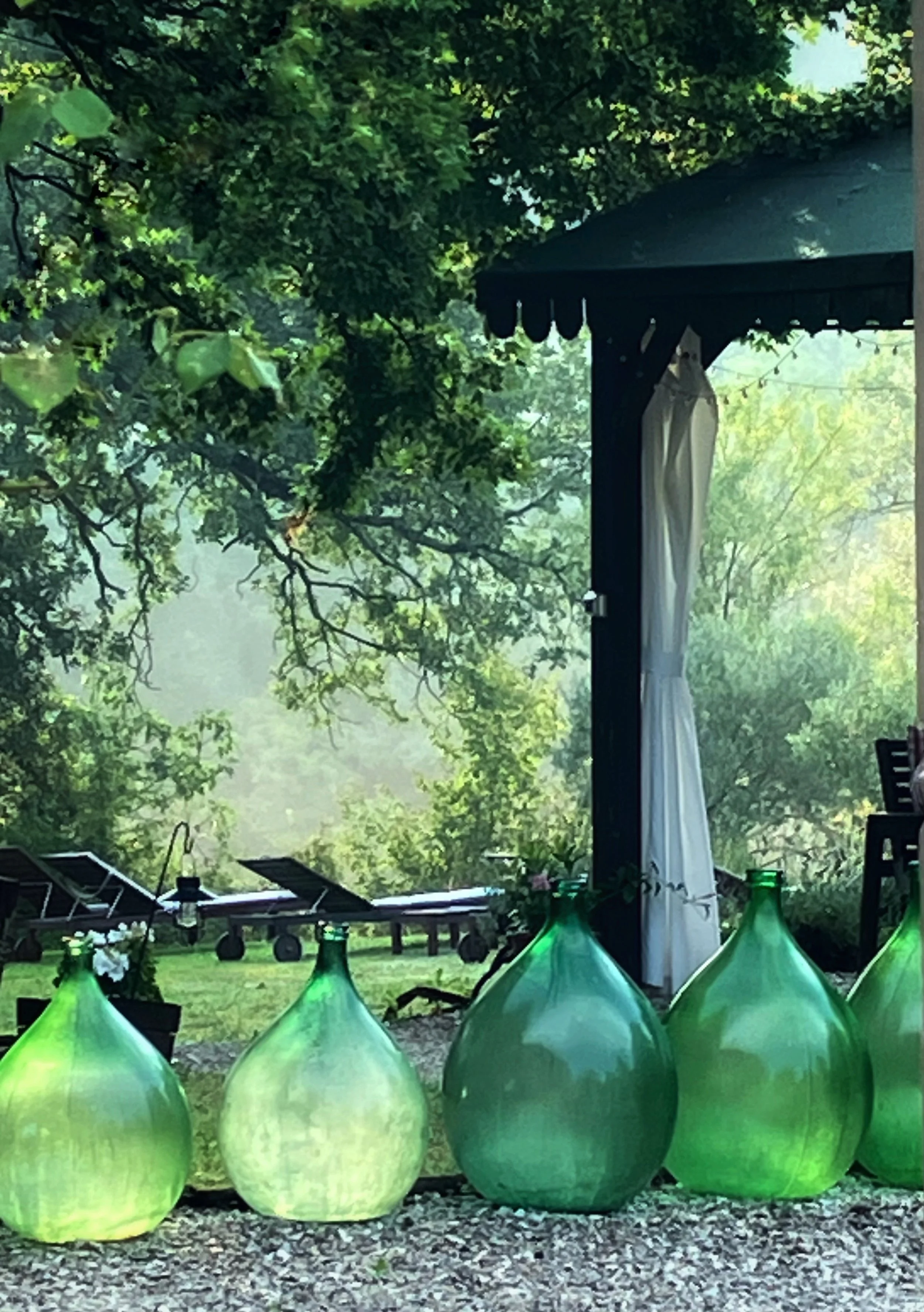 Green glass bottles lined up outdoors on gravel with trees in the background and a wooden structure with white curtains.