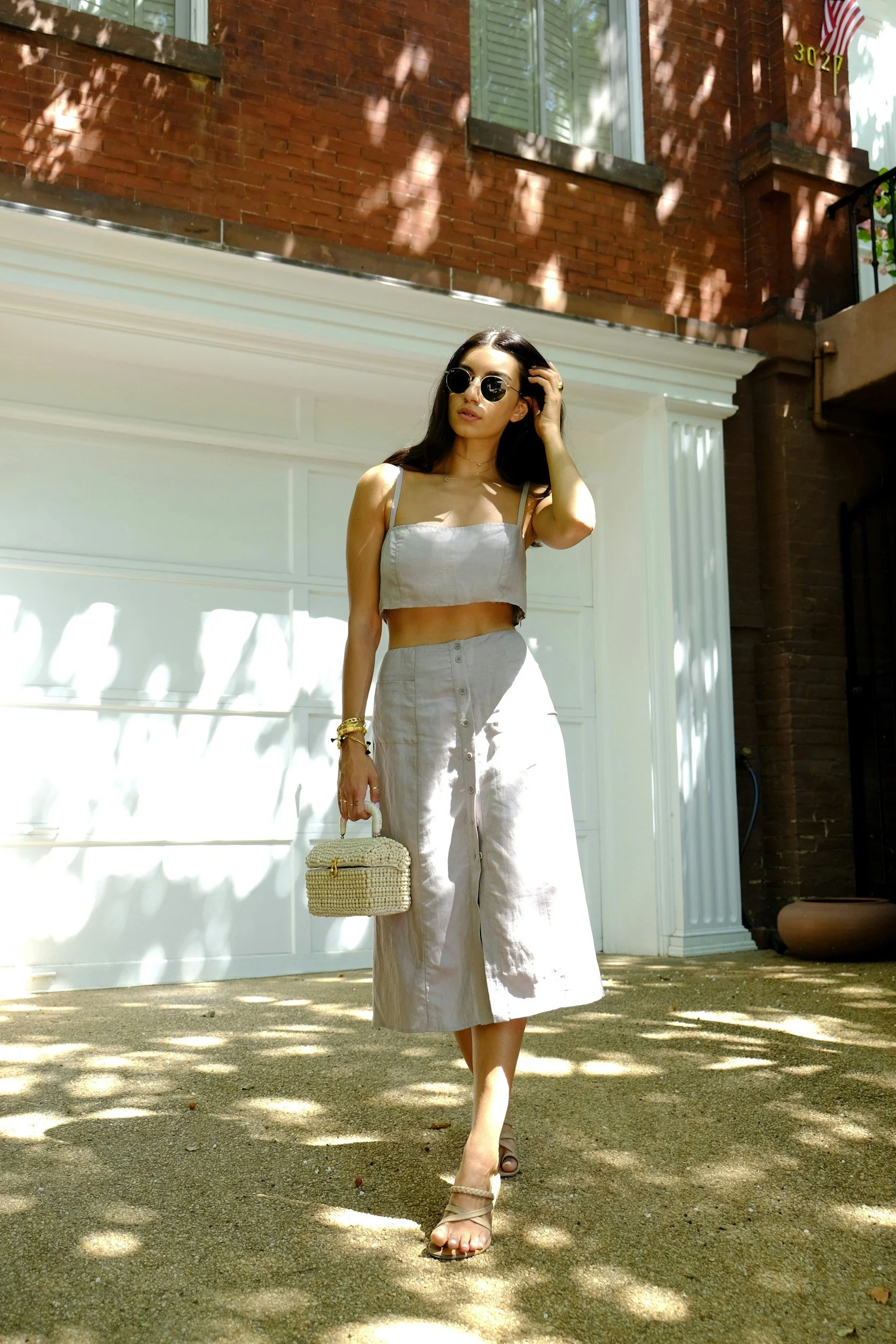 Une femme portant un haut court et une jupe ample, avec des lunettes de soleil, marchant devant une porte de garage blanche, sous un arbre qui projette des ombres sur le sol.