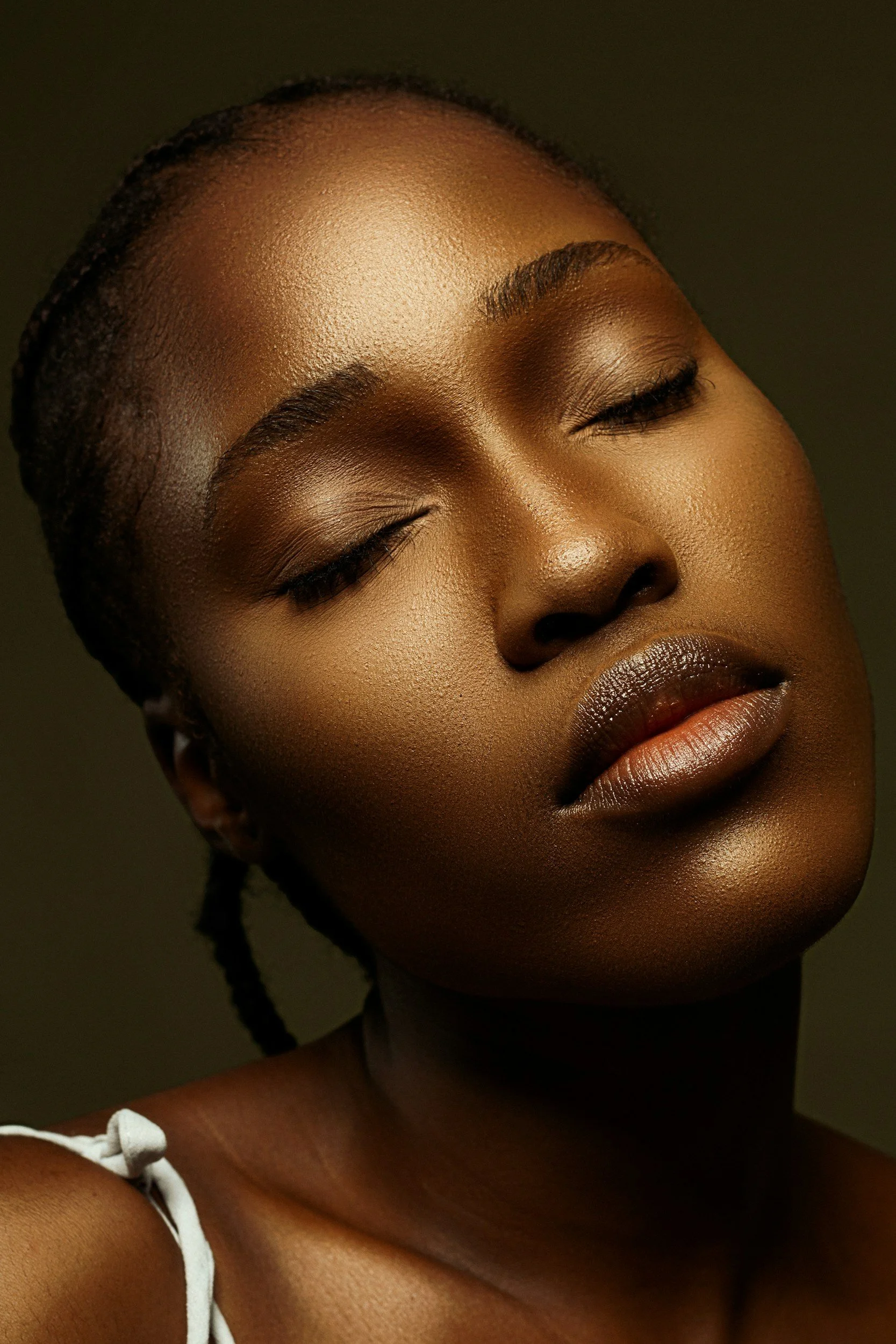 Portrait d'une femme noire aux yeux fermés, peau lisse et maquillage naturel, coiffure en tresses, portant un vêtement blanc à bretelles.