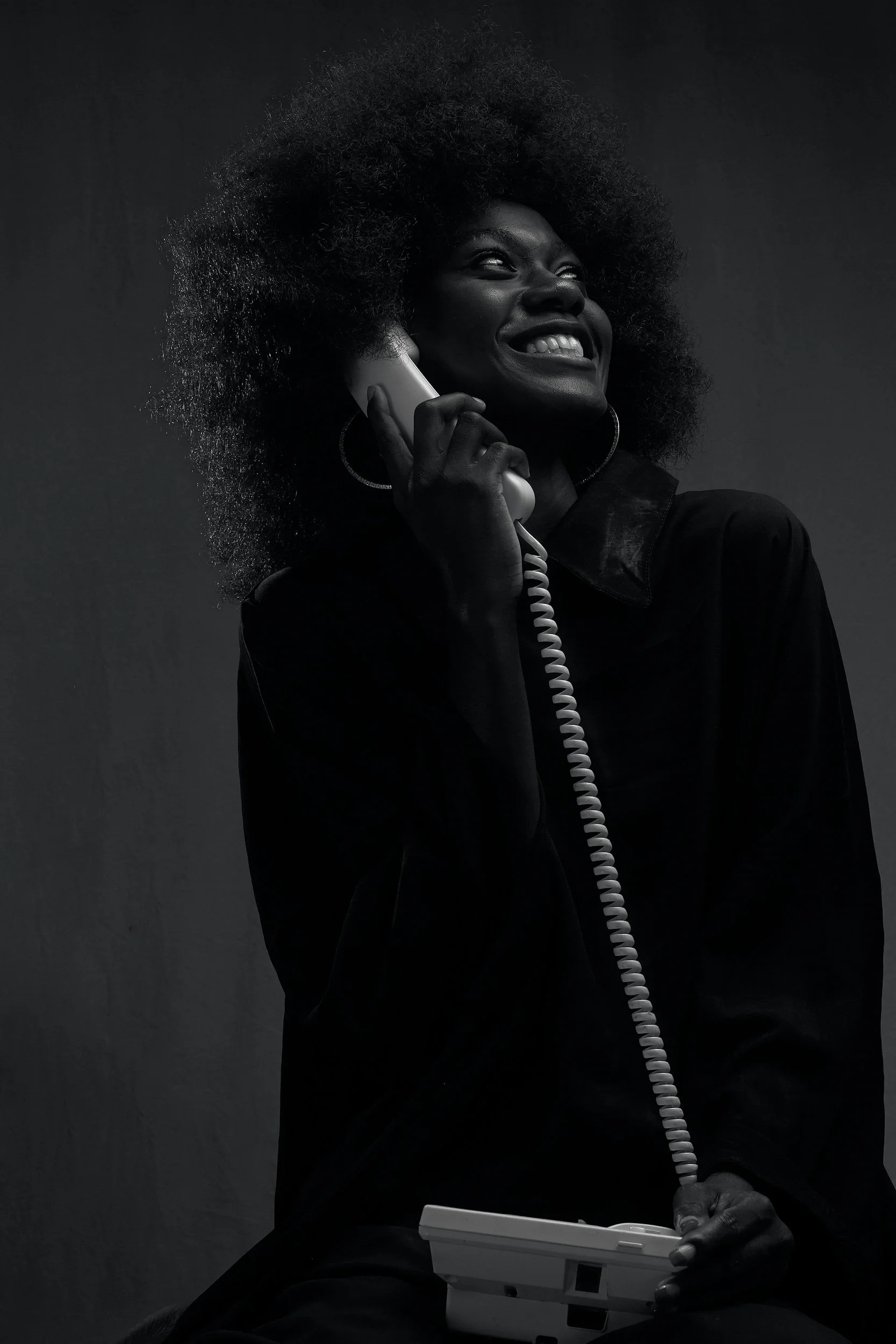Une femme souriante qui parle au téléphone tout en utilisant un appareil électronique, dans un environnement sombre.