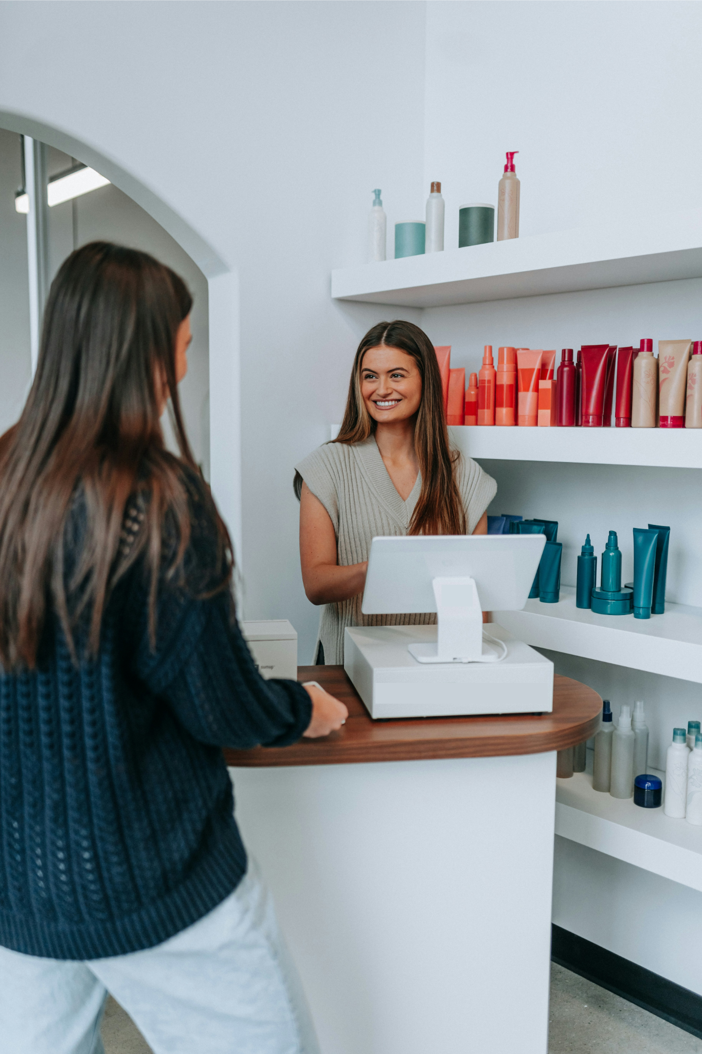 Une vendeuse souriante avec de longs cheveux bruns, portant un haut gris, aidant une cliente dans une boutique de produits de beauté ou de soins capillaires. La cliente, vue de dos, a des cheveux longs et bruns, portant un pull torsadé foncé.