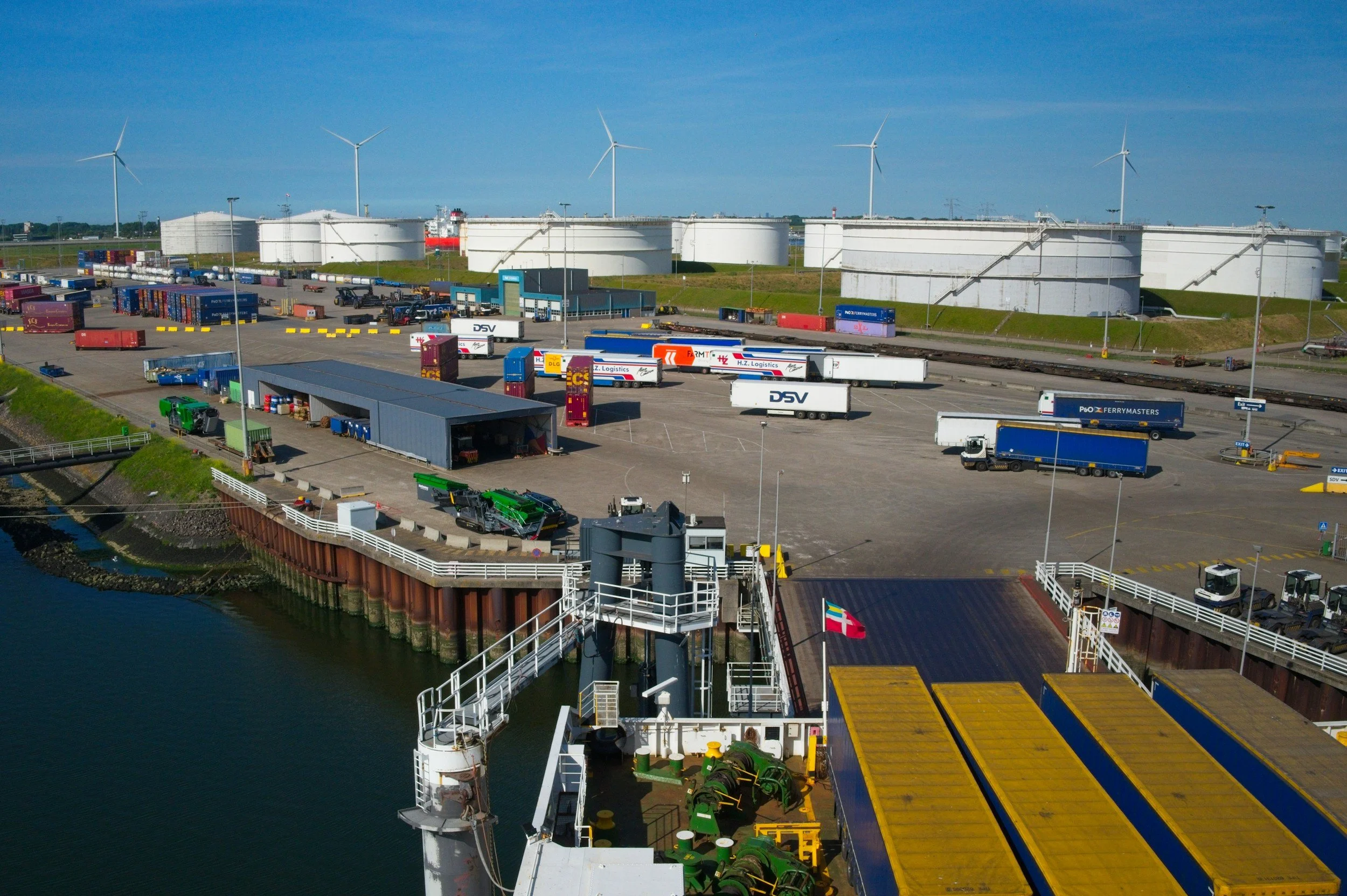 View of a cargo port with multiple shipping containers, trucks, and large white storage tanks, wind turbines in the background, and a body of water in the foreground.