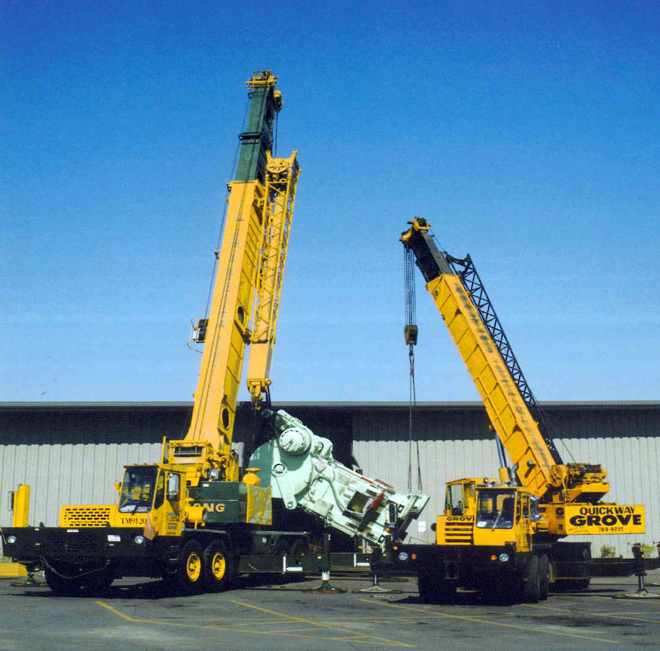 Two yellow mobile cranes with extended booms parked on a paved lot, positioned side by side under a clear blue sky.
