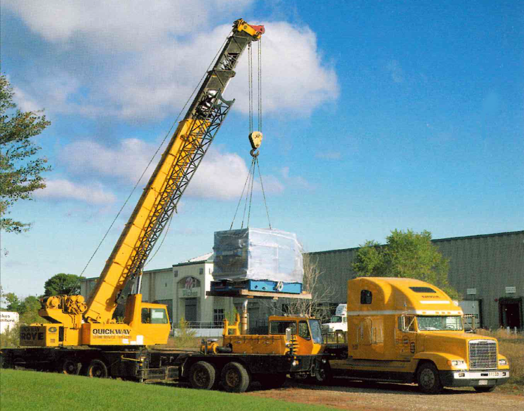 A yellow mobile crane lifting a large container in an outdoor area with a blue sky and some trees and buildings in the background.