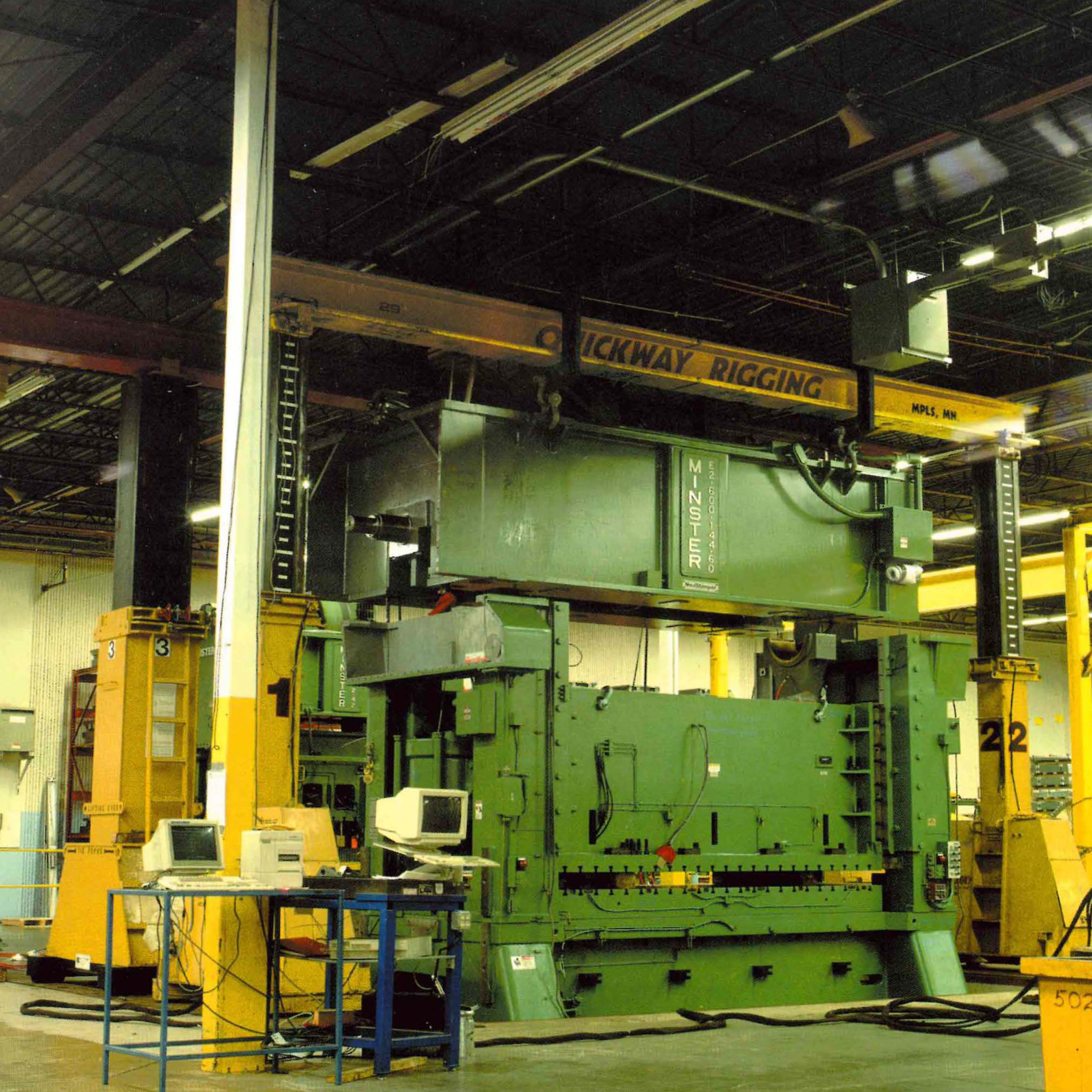 Industrial warehouse with yellow and green machinery, overhead crane labeled "QUICKWAY RIGGING," and various equipment and tools on the floor and shelves.