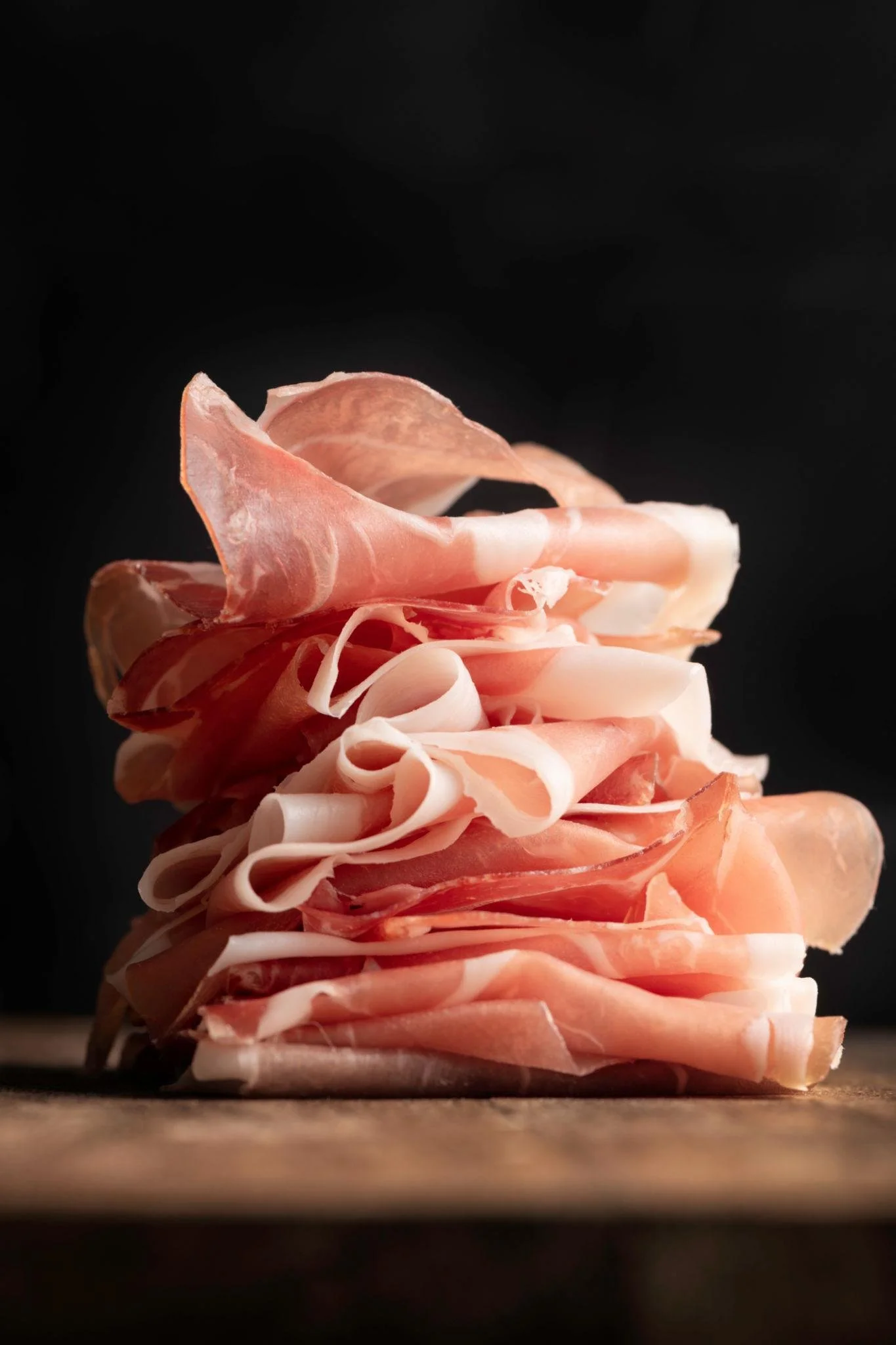 Close-up of sliced prosciutto ham stacked on a wooden surface with a dark black background.