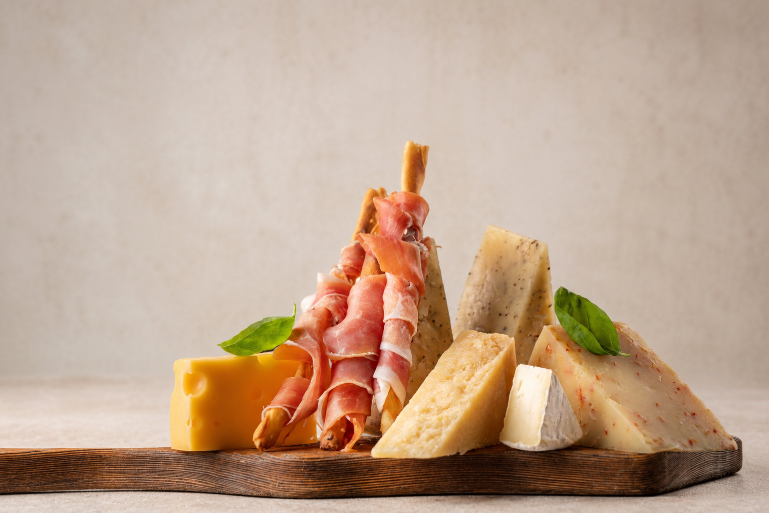 Assorted cheeses and prosciutto on a wooden board with basil leaves.