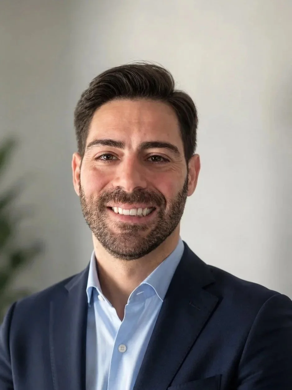 A professional man with dark hair and beard, wearing a dark suit and light blue shirt, smiling at the camera.