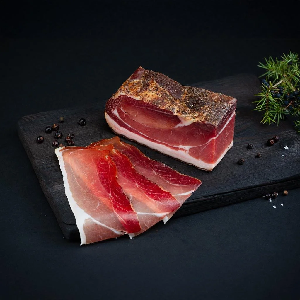 Slices of cured meat, likely prosciutto, on a black cutting board with black peppercorns and some greenery.