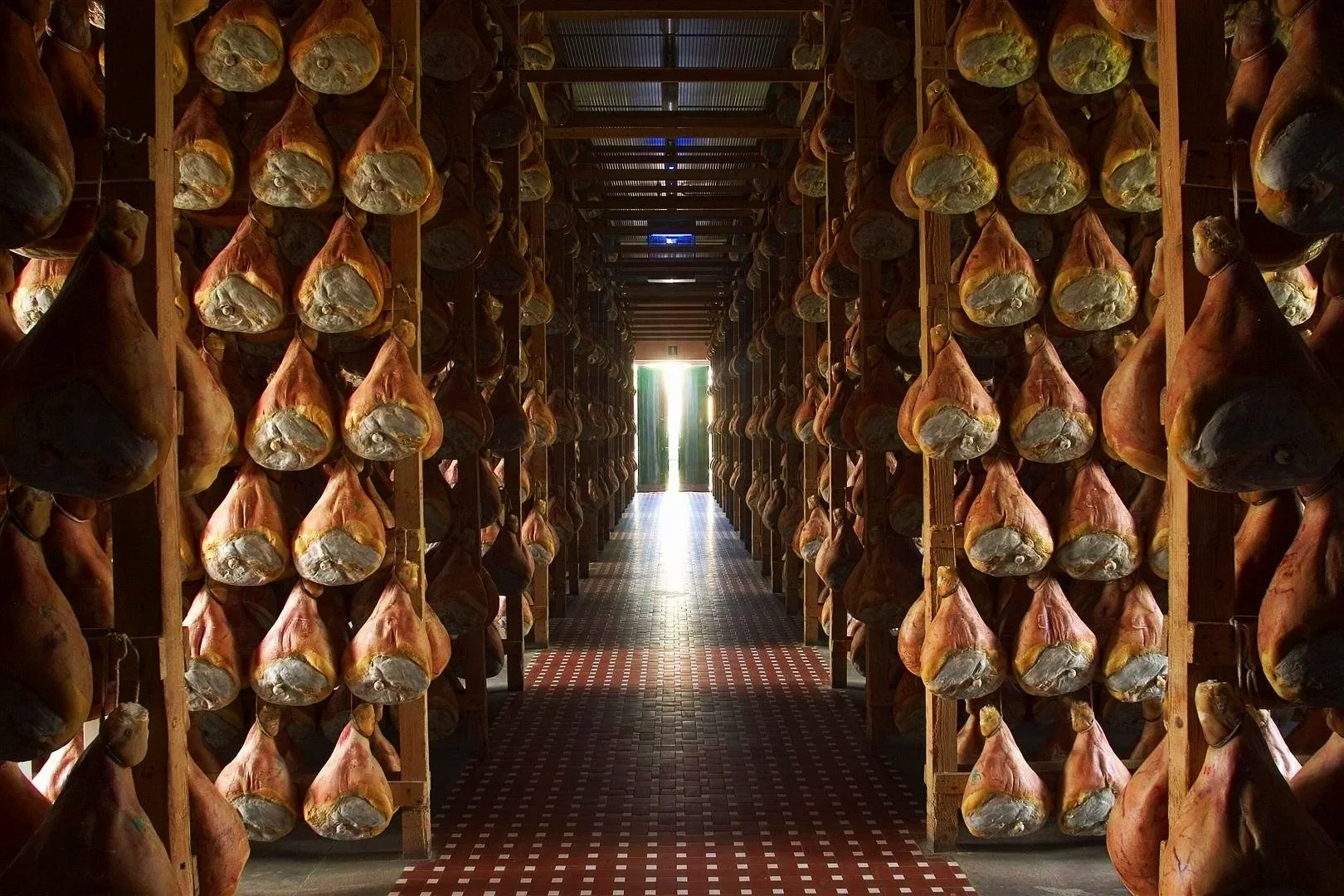 Rows of hanging cured ham legs stored on wooden racks in a curing room with a door at the end allowing light in.