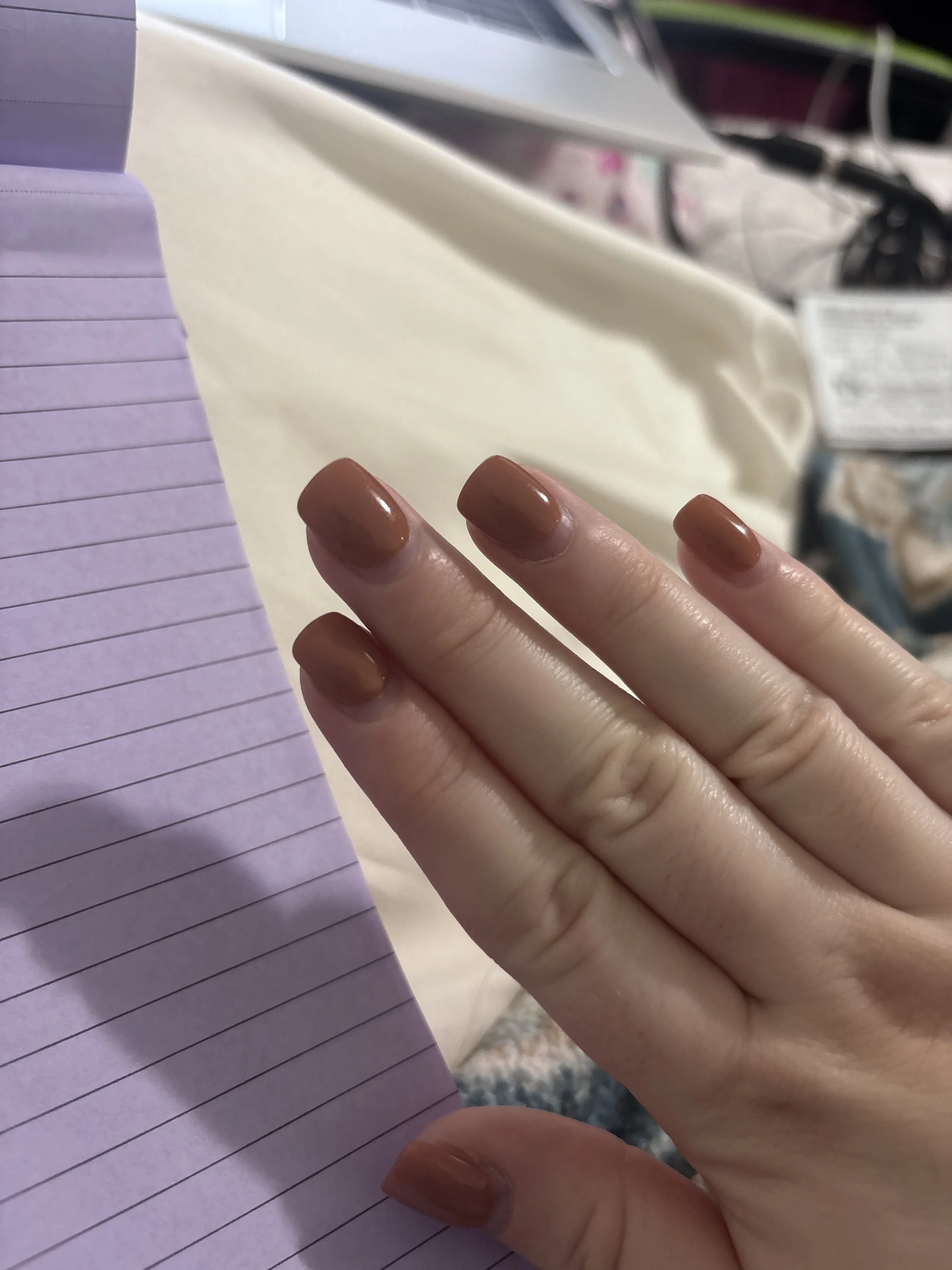 Close-up of a person's hand with tan-colored painted nails resting on a purple lined notebook, with a beige cloth and some electronic cords and devices in the background.