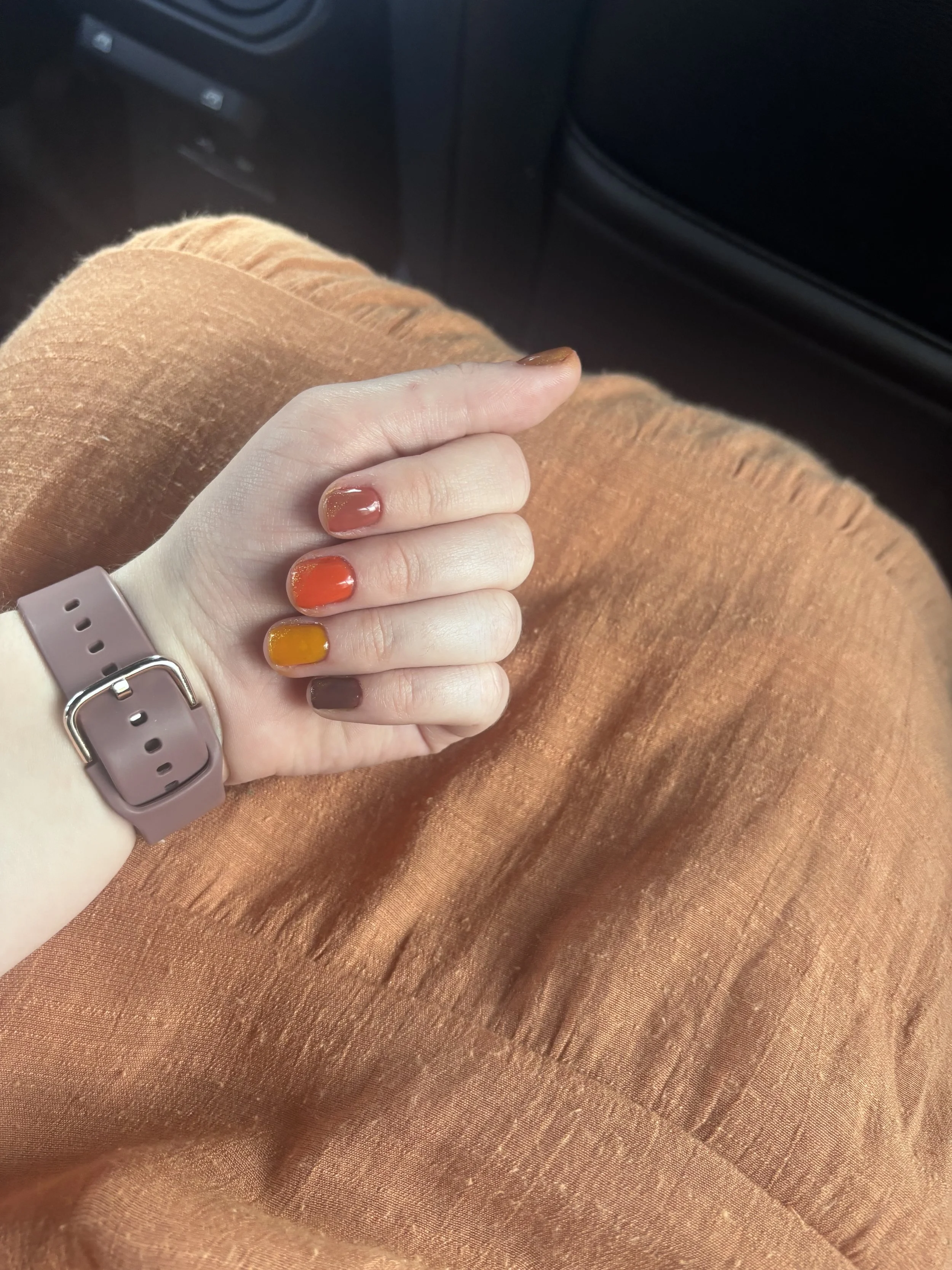 A person's hand resting on a lap with orange and brown painted nails, wearing a pink smartwatch, against orange fabric.