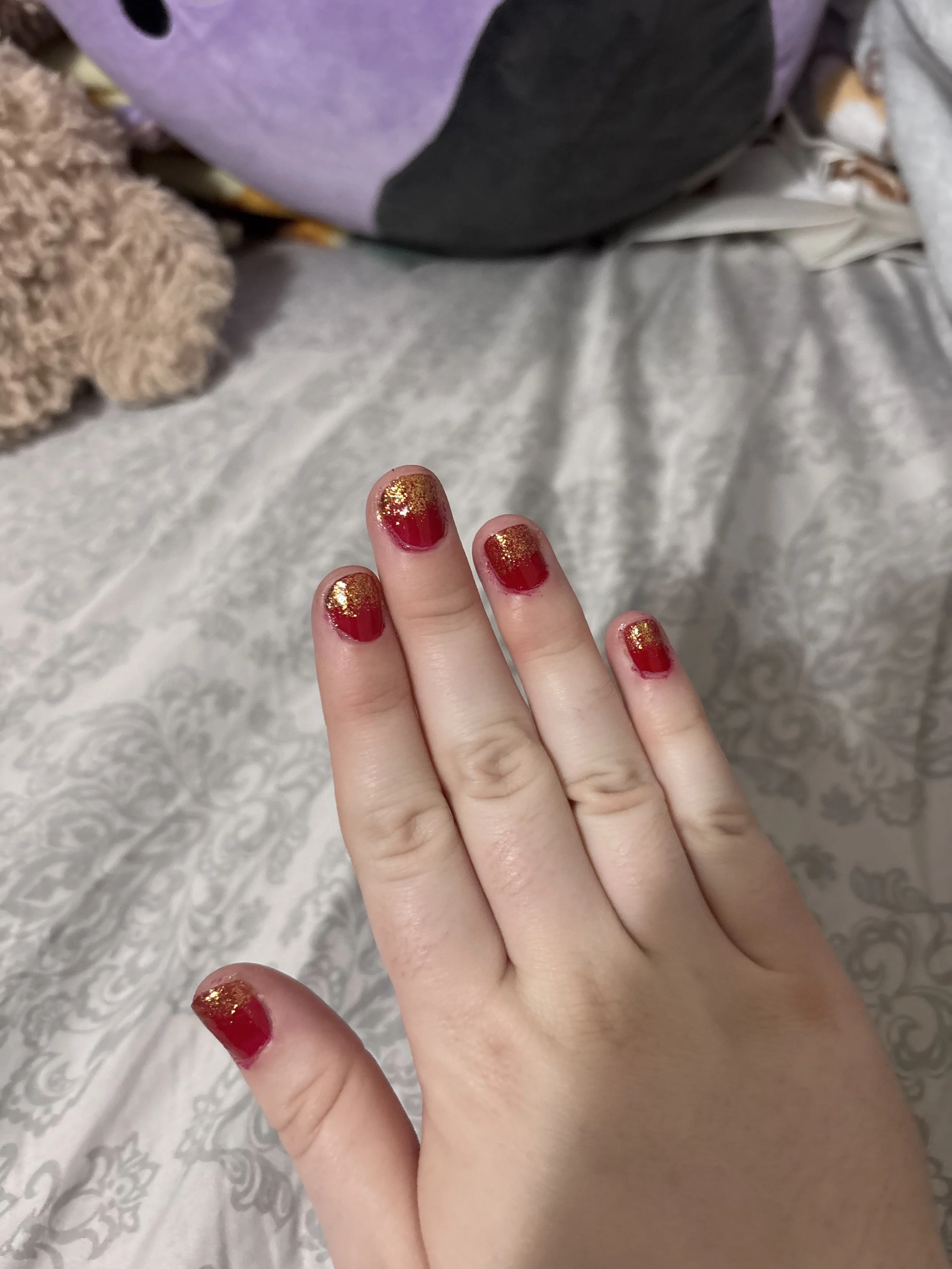 Close-up of fingers with red toenail polish topped with gold glitter, resting on a surface with textured fabric and a purple plush object in the background.