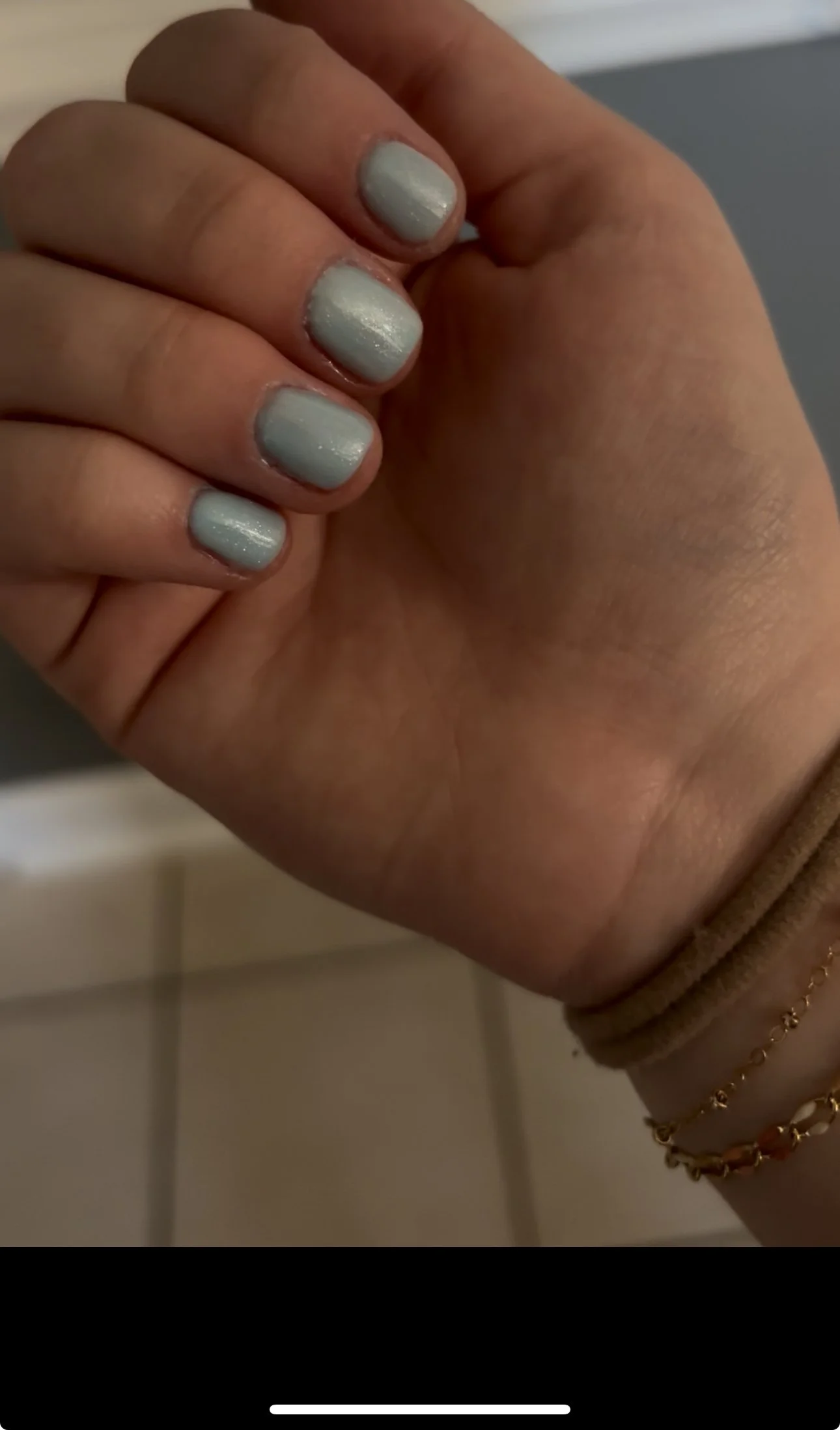Close-up of a hand with neatly painted light gray nails, wearing gold jewelry on the wrist.