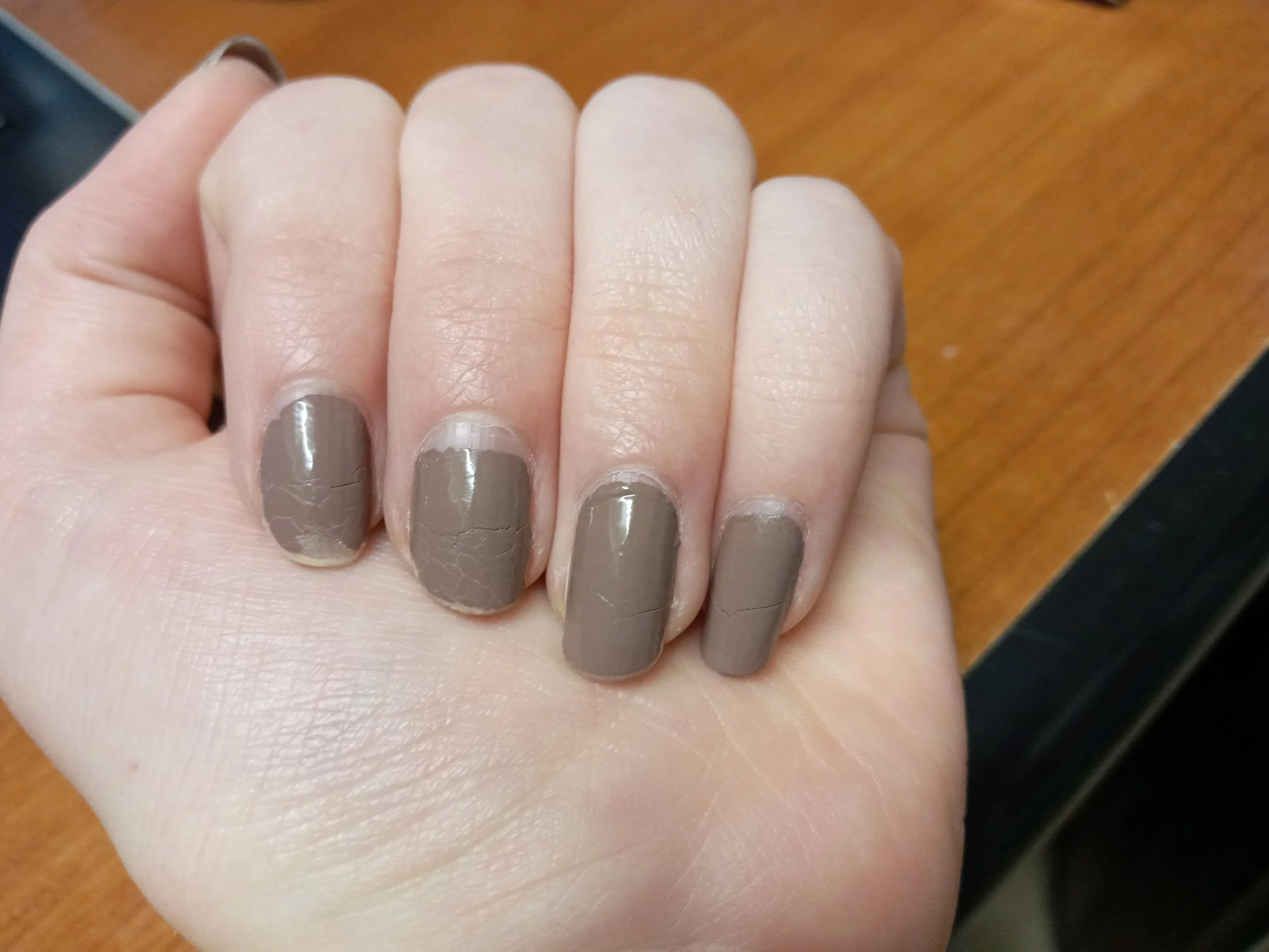 Close-up of a hand with nails painted in a grayish nude color, resting on a wooden surface.