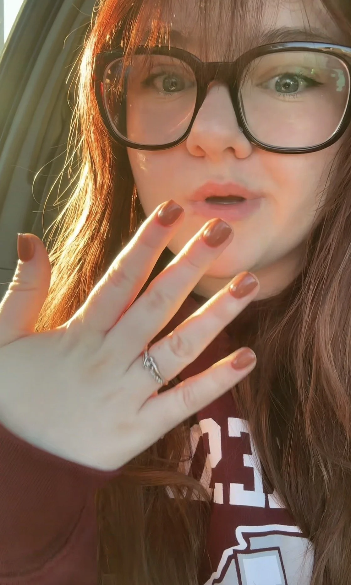 Close-up of a woman with long red hair and large black glasses, showing her face and hand with mauve nail polish, an engagement ring, and a maroon hoodie, sunlight illuminating her hair.