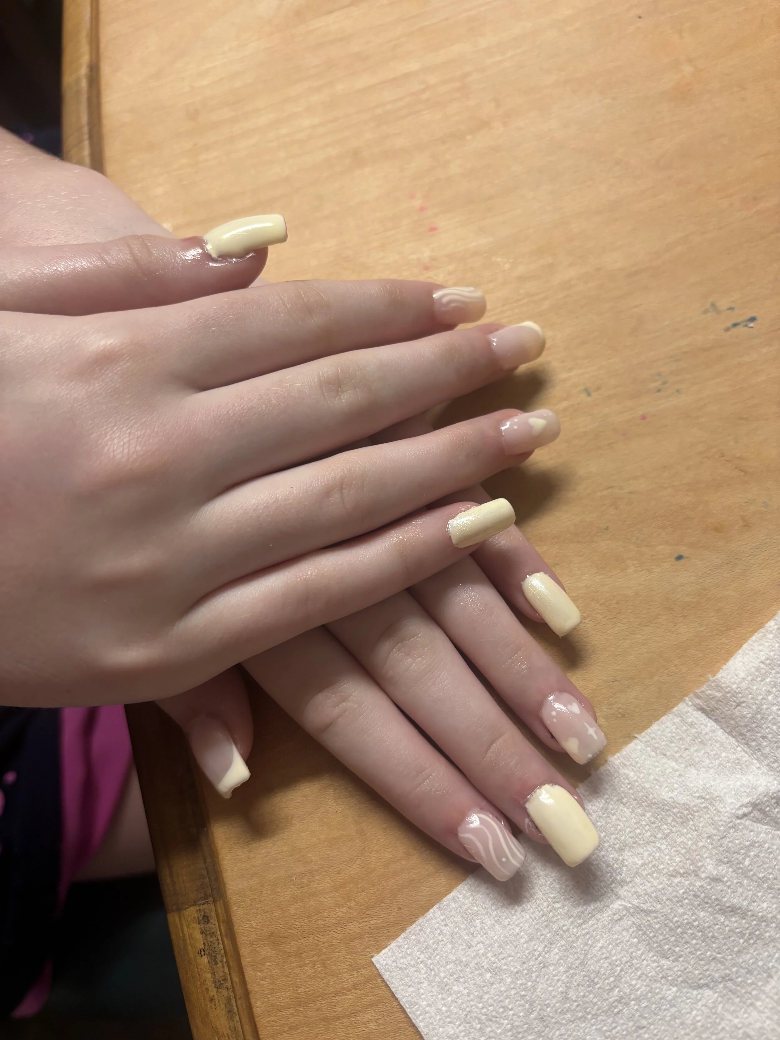Close-up of a person's hand with freshly painted nails in a light cream color, lying on a wooden surface with some small paint smudges, and a paper towel underneath.