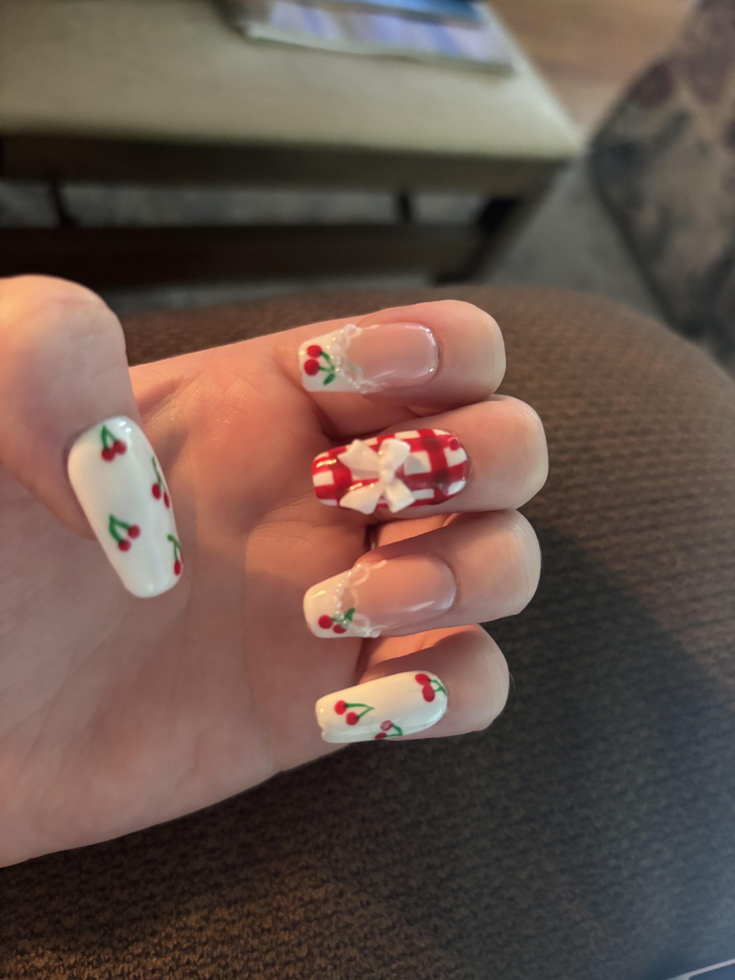 Hand with decorated Christmas-themed nails displaying small red holly berries and green leaves, with a red and white checkered pattern on the ring finger, held up over a dark surface.