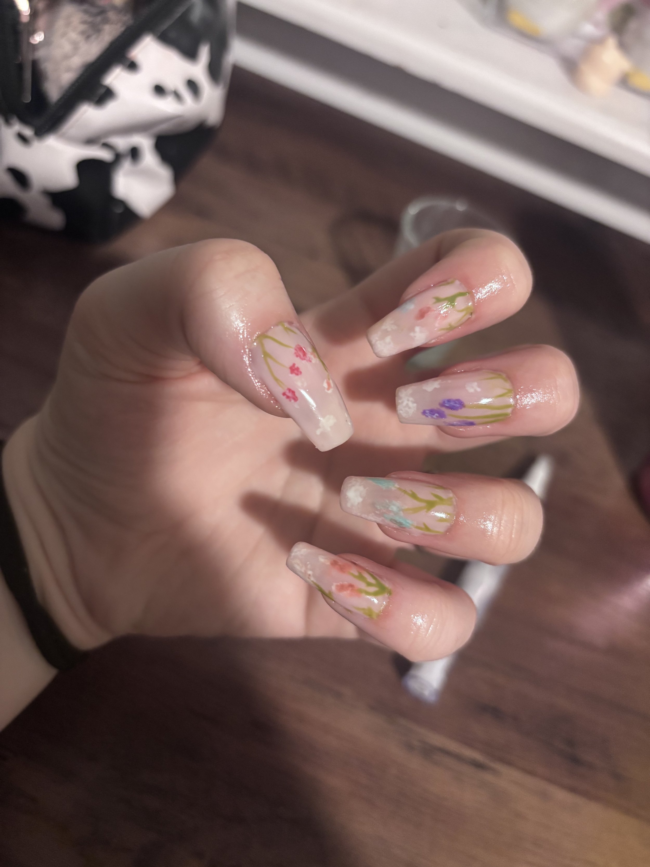Close-up of a hand with decorated, long acrylic nails featuring floral designs, with a blurred background.