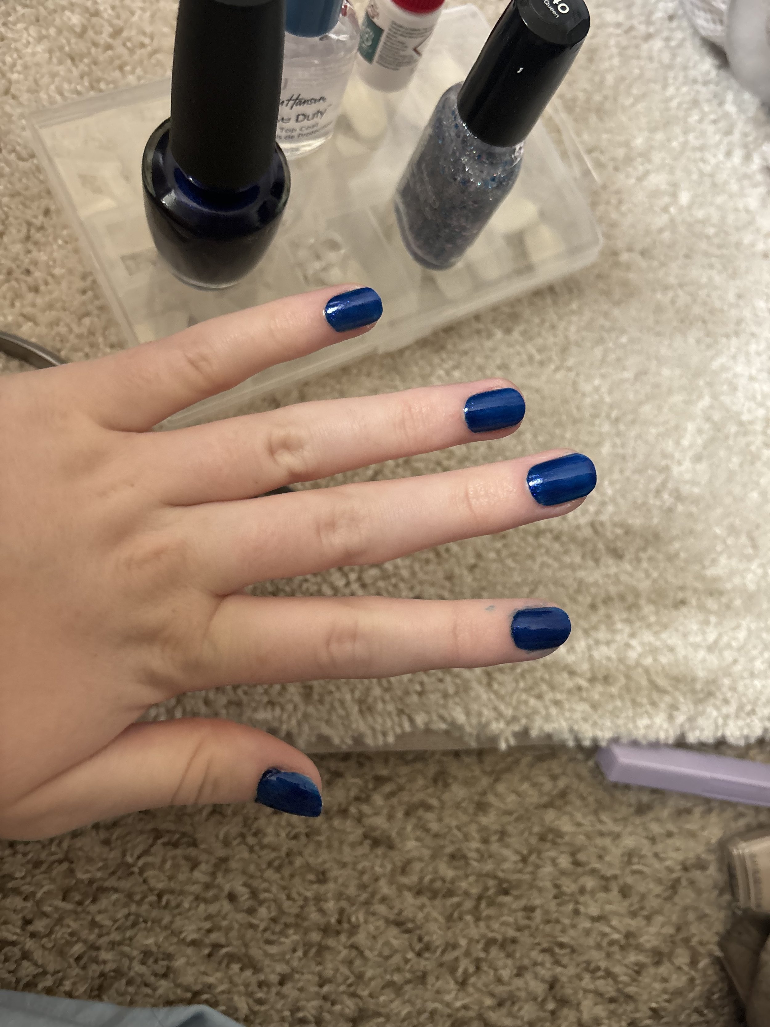 A person's hand with nails painted blue is shown in the foreground. In the background, there is a clear plastic nail polish organizer with several bottles of nail polish, glitter, and other nail accessories on a beige carpet.