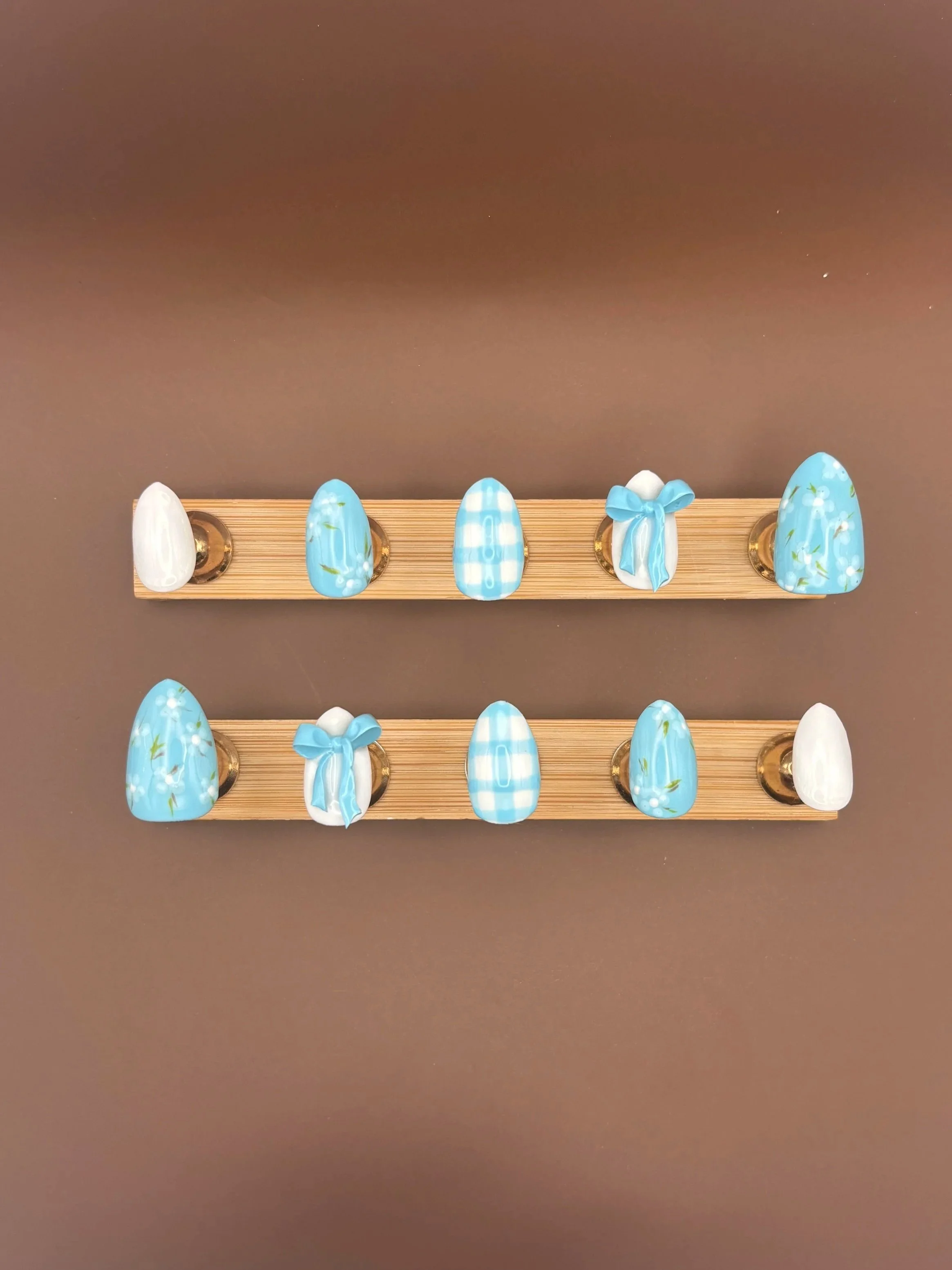 Two wooden display racks holding decorated Easter eggs with blue, white, and green floral and checkered patterns and bows, placed on a dark surface.