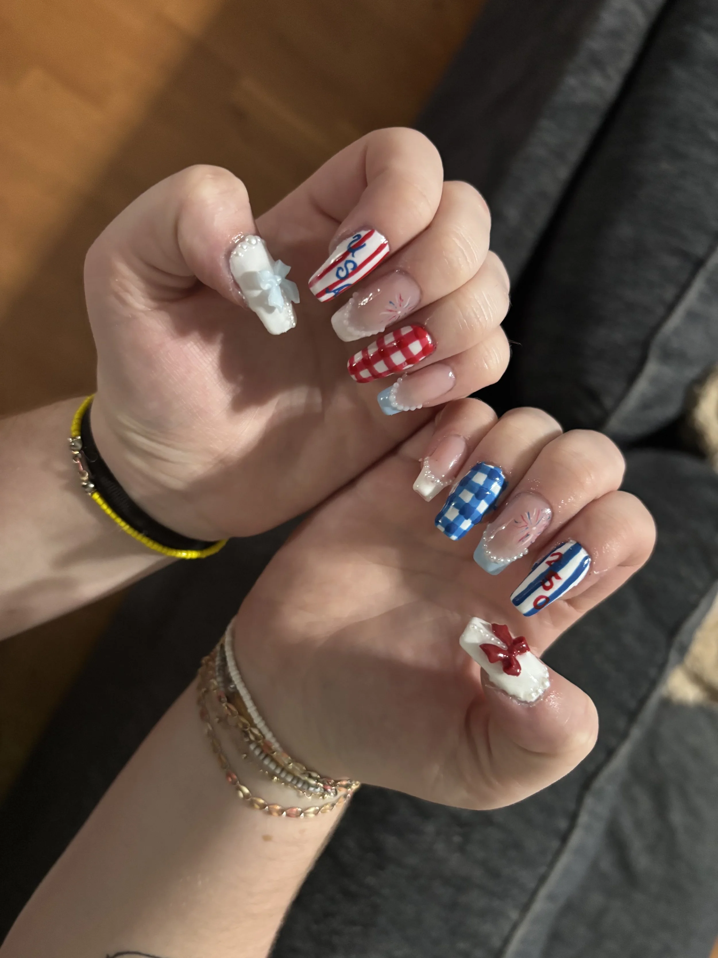 Close-up of a person's hand with a festive manicure featuring patriotic-themed nail art including red, white, and blue stripes, checks, and embellishments, each nail with a different design.