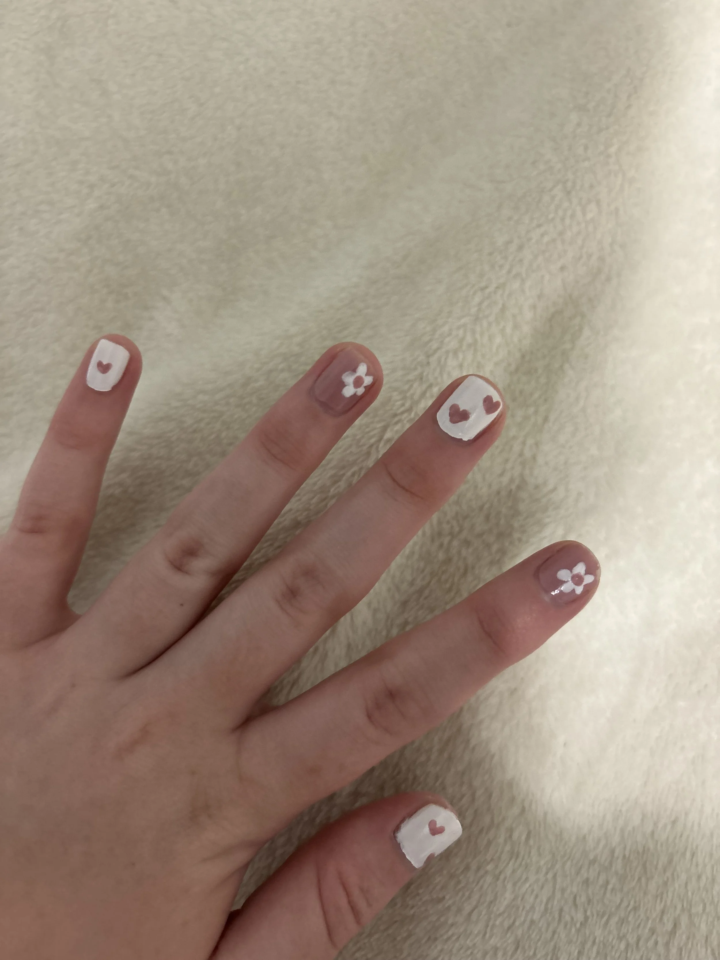 Close-up of someone's hand showing painted nails with white backgrounds and small red hearts or white flowers on each nail, against a beige textured surface.