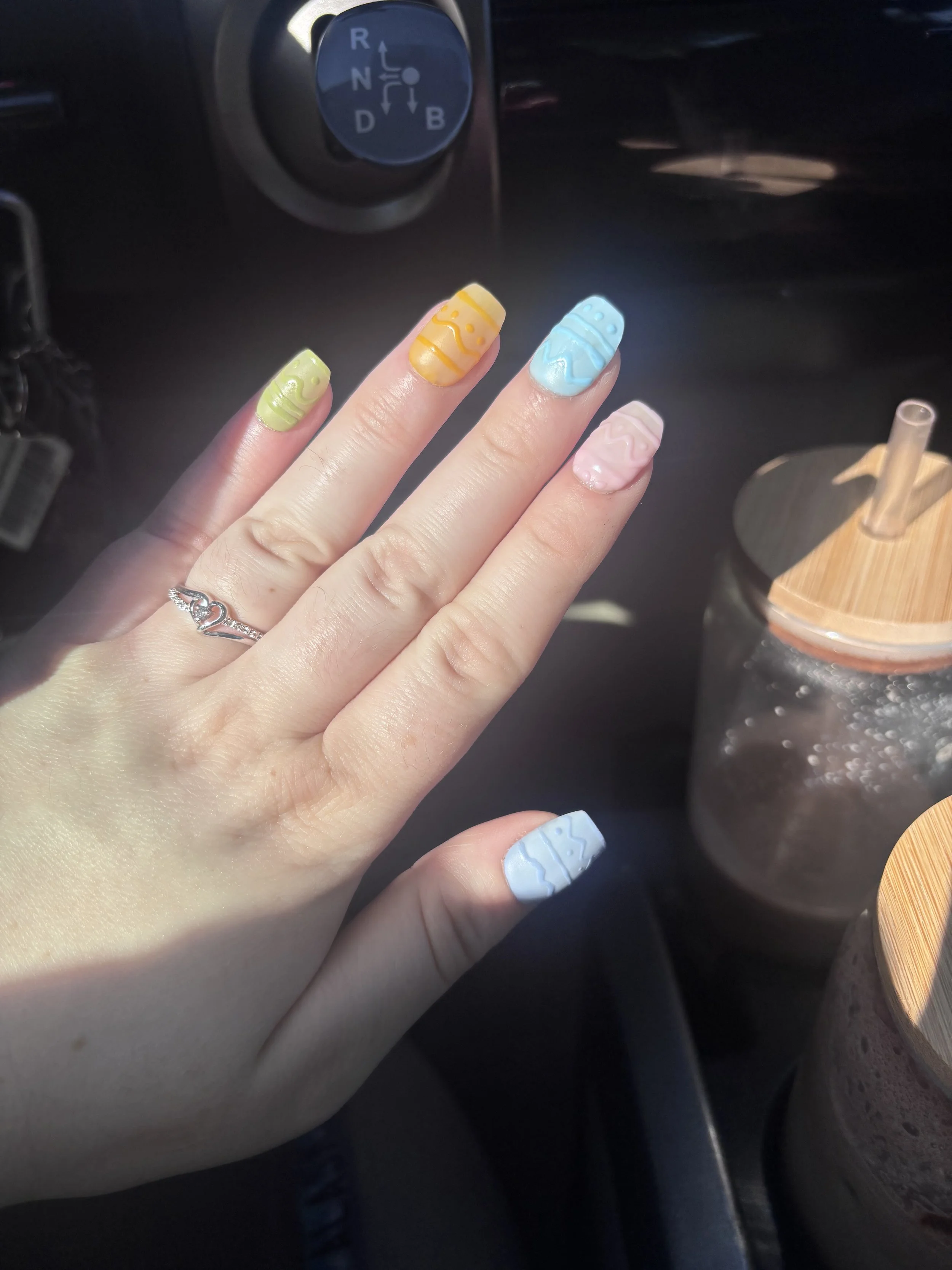 Hand with pastel-colored, patterned nails in green, yellow, blue, and pink, resting on a car's steering wheel with a gear shift indicator in the background.