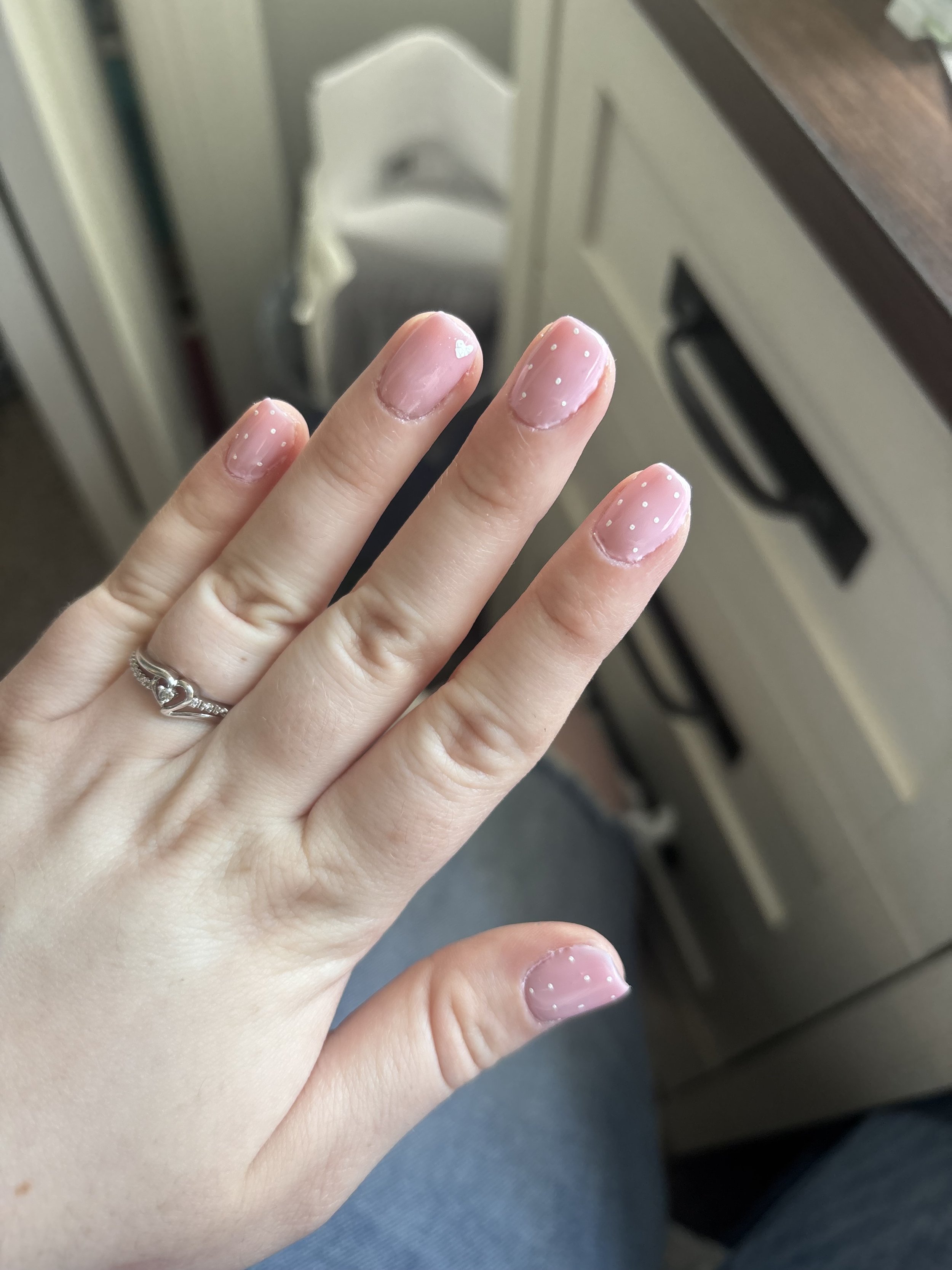 Close-up of a person's hand with decorated nails, showing four nails with pink nail polish and small white dots, and a silver ring on the ring finger. The hand is held up against a kitchen environment with a white stove or oven in the background.