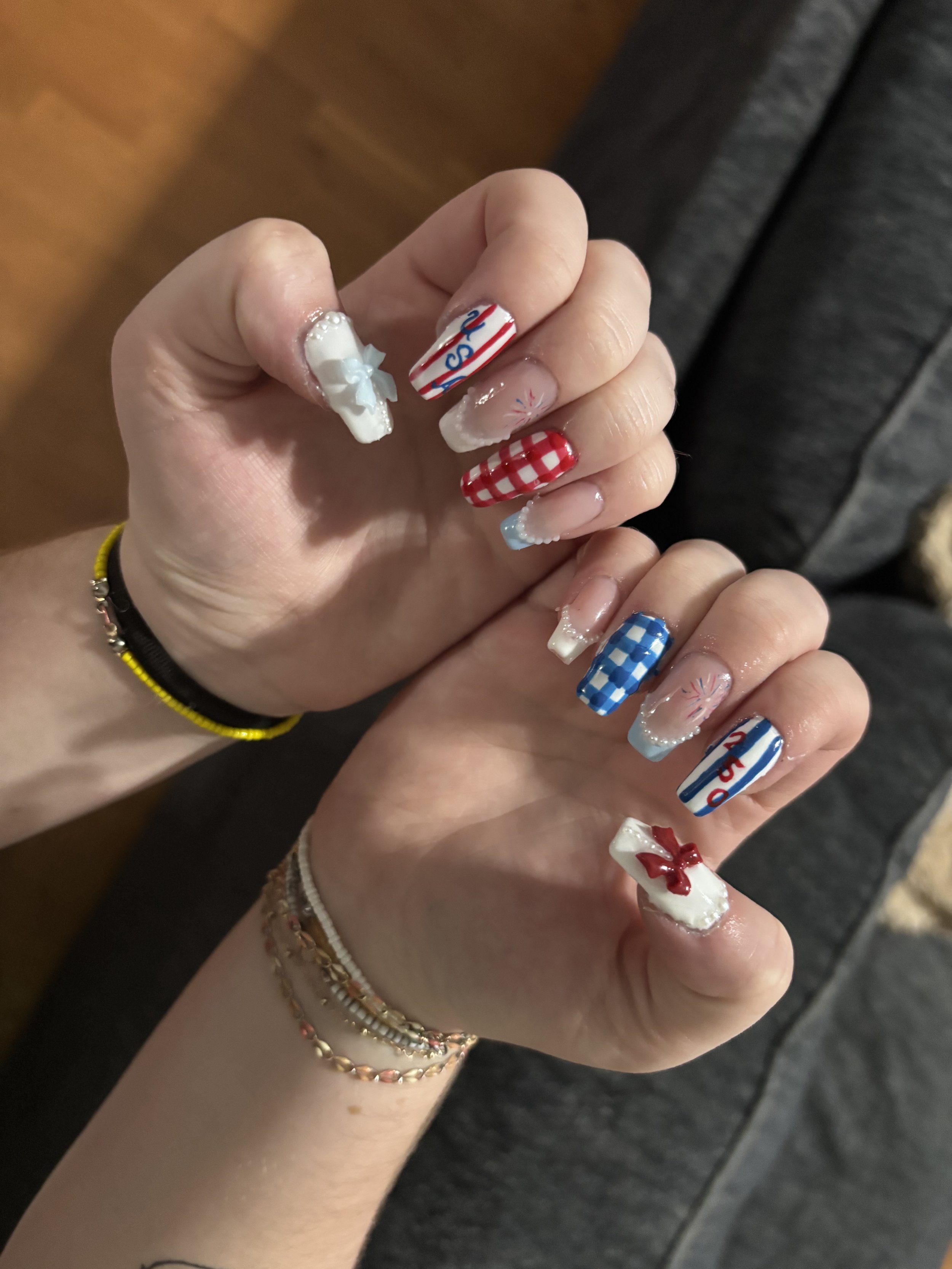 Close-up of a hand with decorated nails featuring patriotic and holiday-themed designs, including a red and white checkered pattern, a white ribbon bow, and festive slogans.