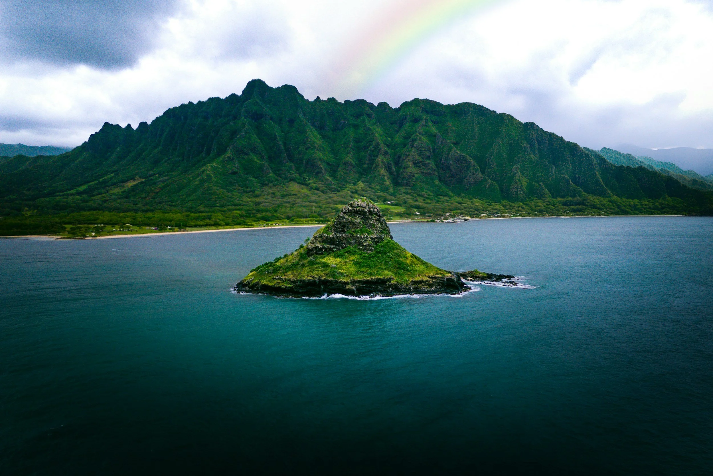 Lush Hawaiian mountains and a small island surrounded by turquoise water with a rainbow in the distance.