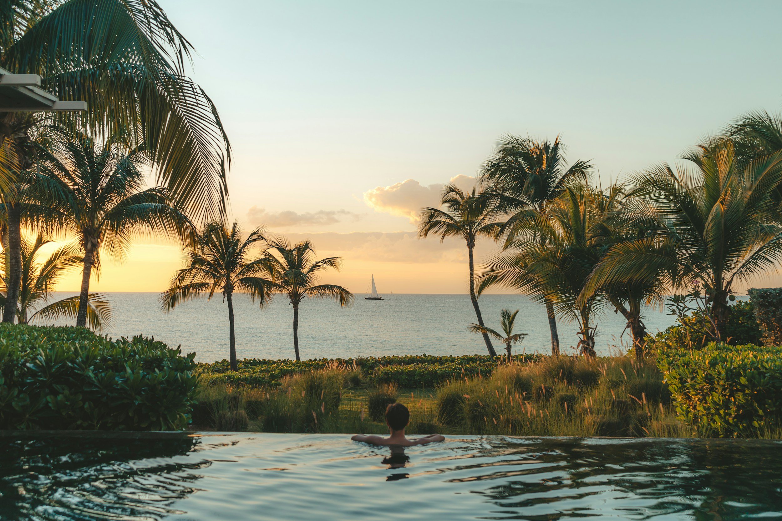Traveler relaxing in an infinity pool overlooking a tropical beach with palm trees and sailboats at sunset.