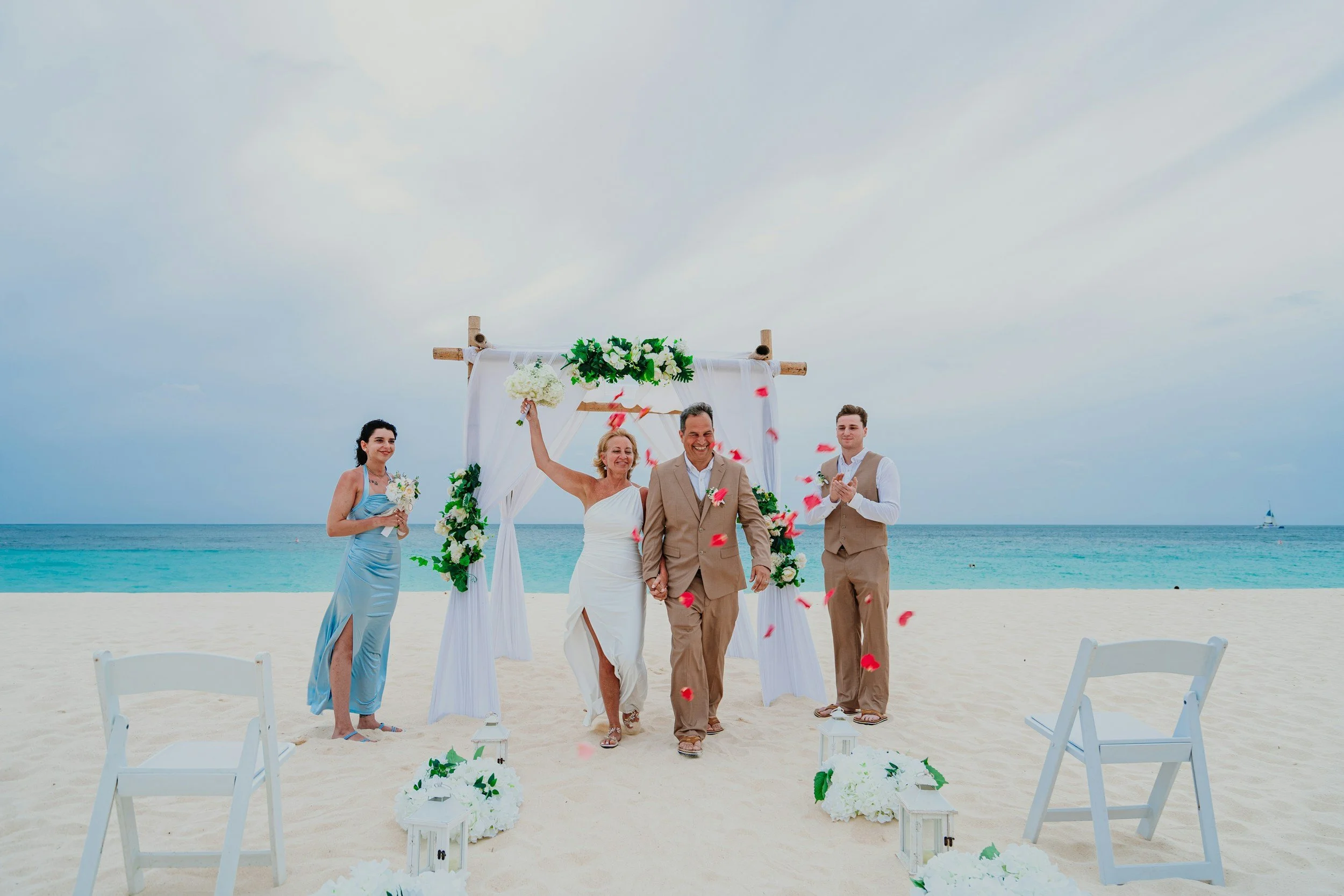 A wedding ceremony on the beach with a decorated arch, the bride and groom holding hands and smiling, surrounded by bridesmaids and groomsmen, with chairs and floral arrangements in the foreground and the ocean and sky in the background.