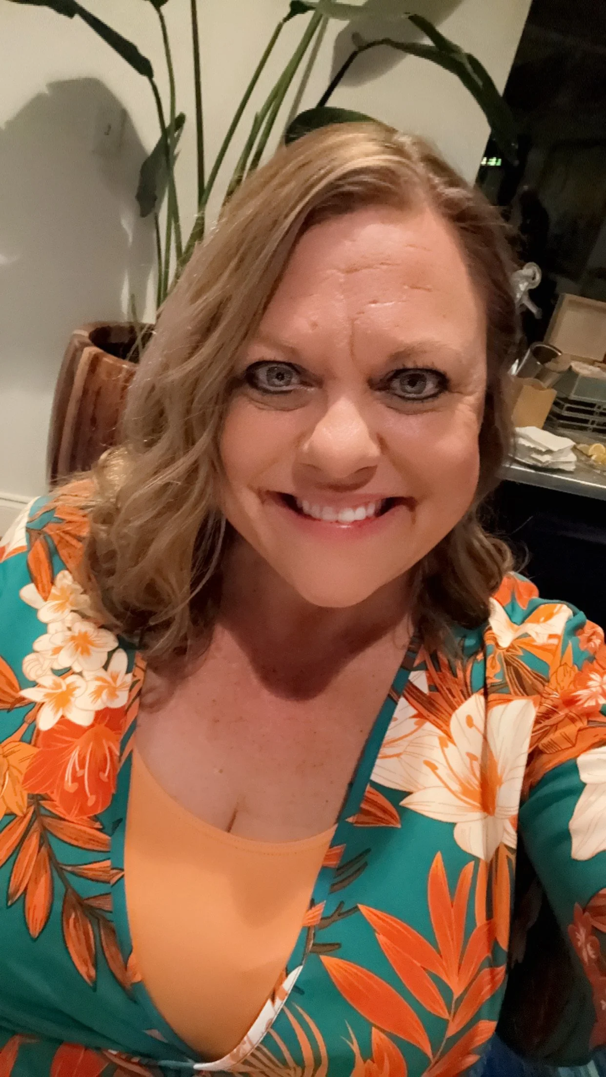 A woman with wavy light brown hair and blue eyes smiling for a selfie. She is wearing a colorful blouse with a floral and leaf pattern in orange, white, and teal. In the background, there is a large potted plant with long green leaves, a white wall, and some furniture with dishes and napkins.