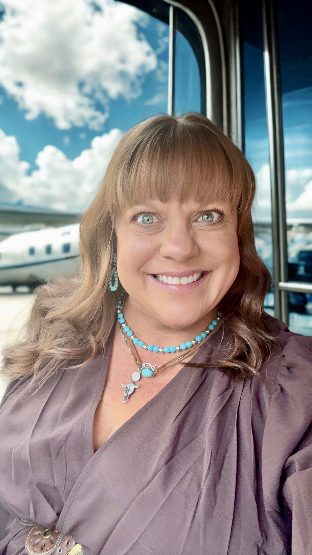 A woman smiling with blue eyes and brown hair, wearing turquoise jewelry, standing inside an airport with a backdrop of planes and windows showing a cloudy sky.