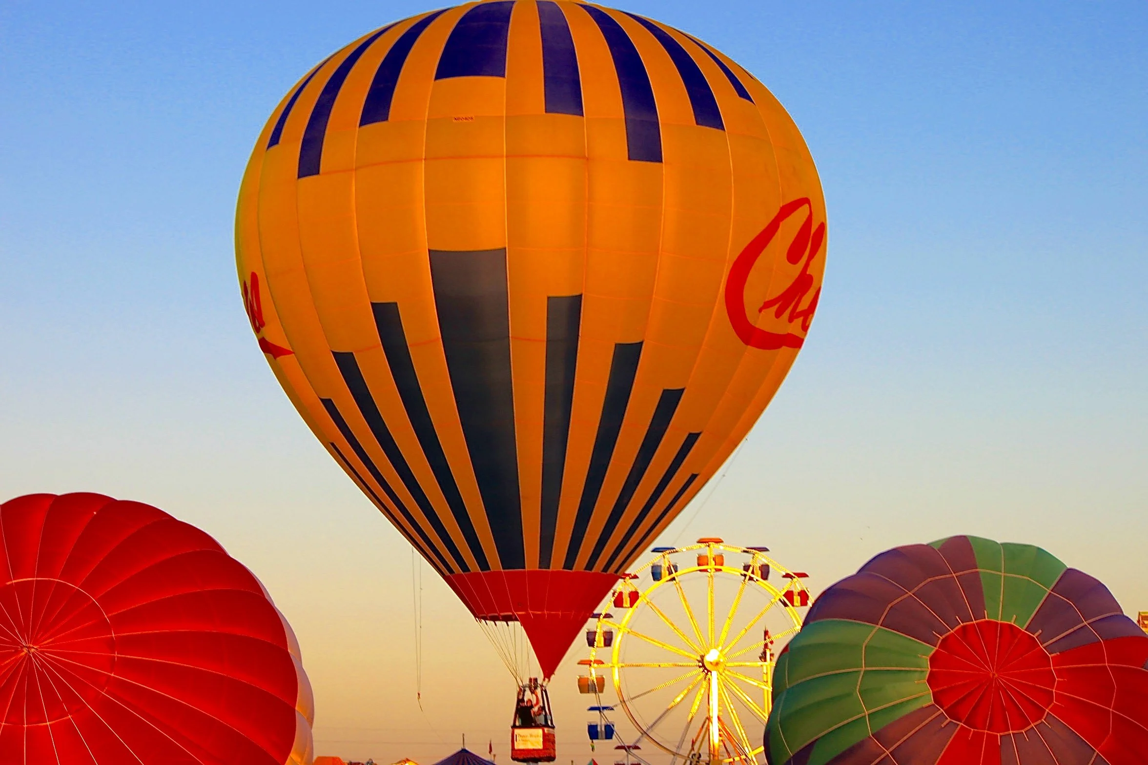 Several colorful hot air balloons floating in the sky, including a prominent large orange and black striped balloon and others in red, green, purple, and yellow, with a Ferris wheel visible below them at a festival or fair during a sunset.