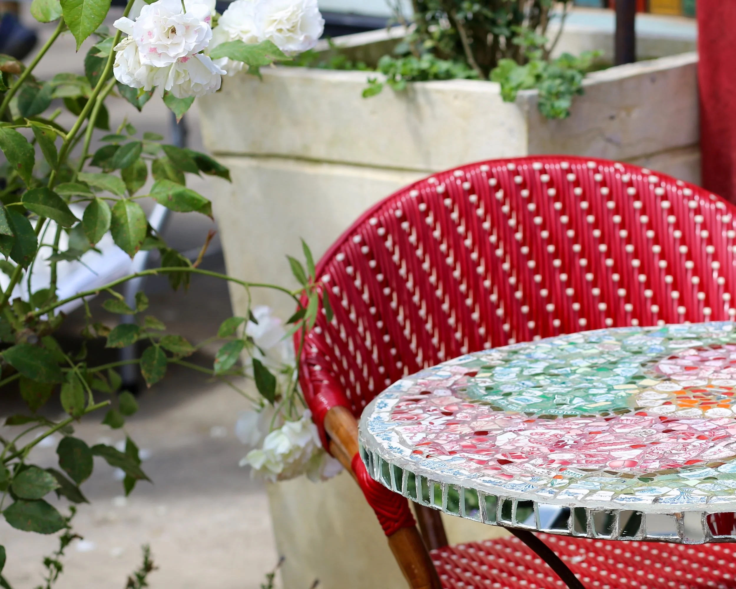 A round table covered with a colorful mosaic tabletop, a red woven chair partly visible, and a large potted plant with white flowers and green leaves in the background. A bistro table at a cafe in St. Helena CA