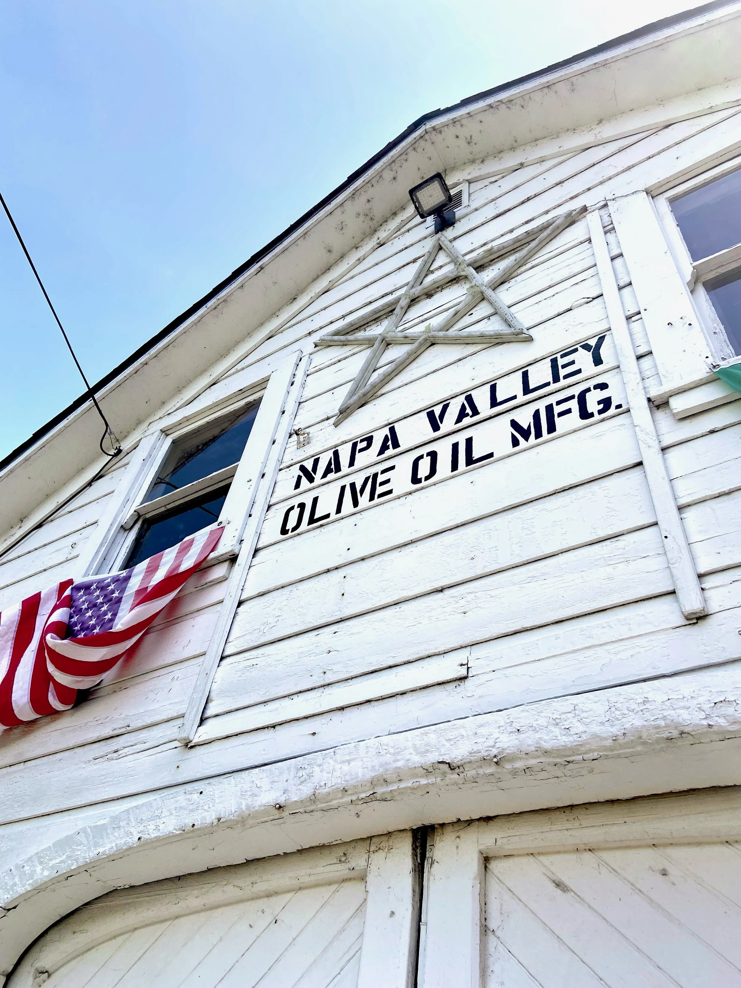 The side of an old white wooden house with a window, a star decoration above the window, a sign with the words "Napali Valley Olive Oil Mfg." and American flags hanging below.