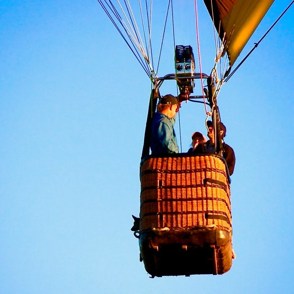 Hot air balloon basket in the sky with three men inside, surrounded by colorful ropes and fabric, against a blue sky, flying over Oakville CA