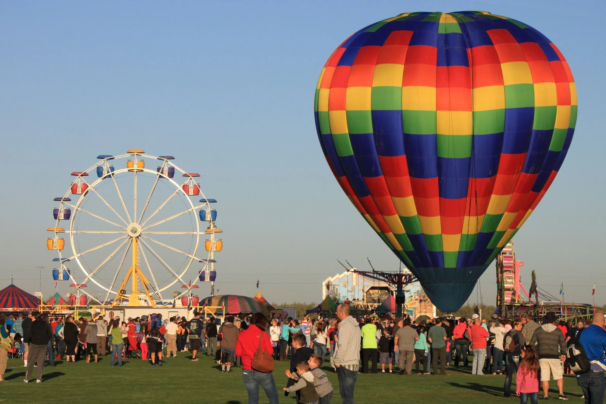 A colorful hot air balloon with a checkered pattern of red, yellow, green, and blue is floating in the sky at a fairground filled with people. In the background, there is a Ferris wheel and other amusement, Launching from Rutherford CA