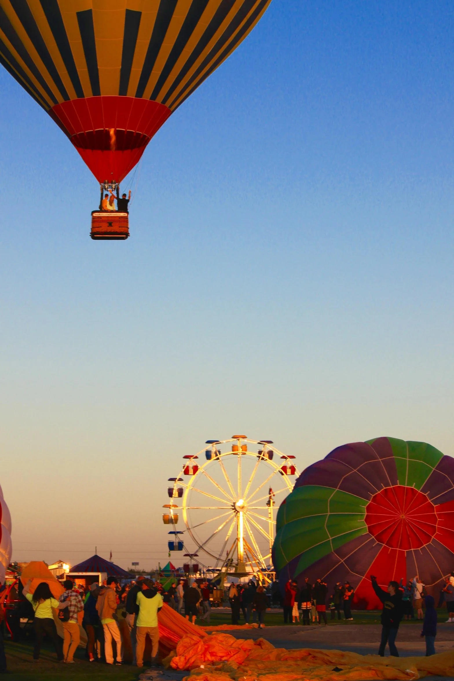 A large hot air balloon is being inflated at a fairground during sunset, with a crowd of people watching. In the background, there are a Ferris wheel and other fair rides.