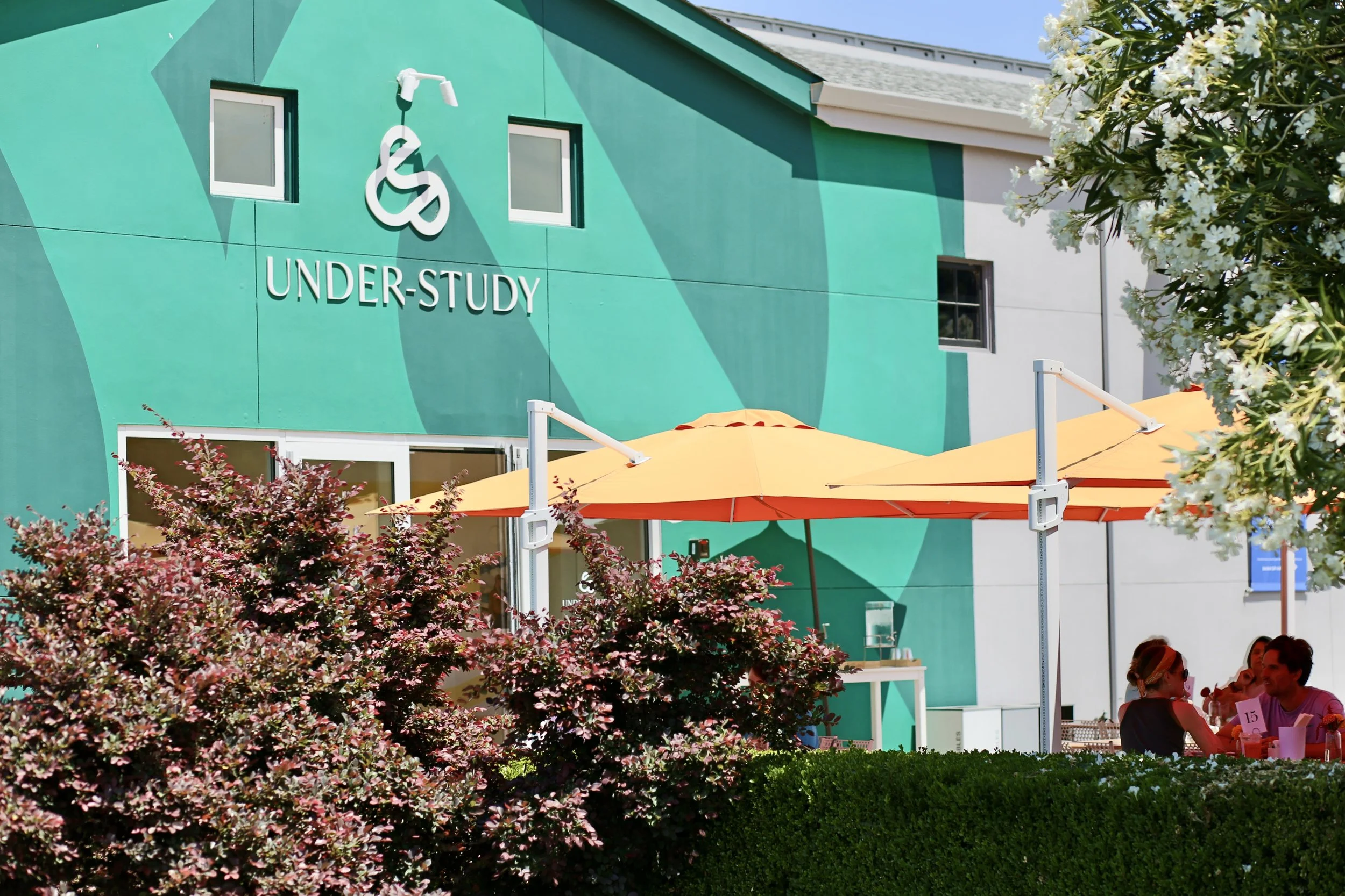 An outdoor scene at a shopping center or mall with a turquoise building that has the words 'UNDER-STUDY' on it. There are trees with white and pink flowers, and yellow umbrellas shading outdoor seating where people are sitting.
