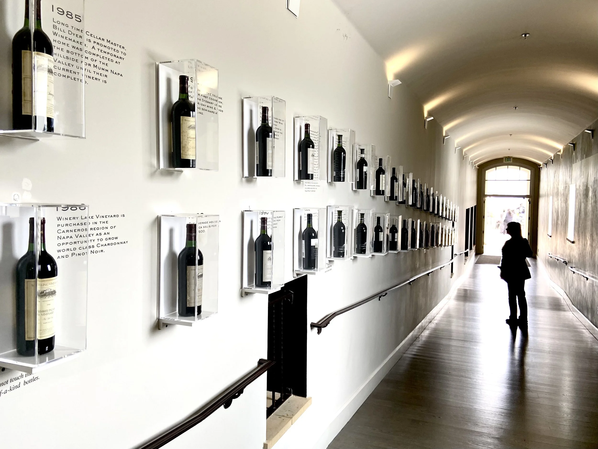 A hallway with a white wall displaying several bottles of wine in clear display cases and informational plaques. The hallway has a curved ceiling with upward lighting fixtures and a door at the end with a window allowing light to come through. A pers