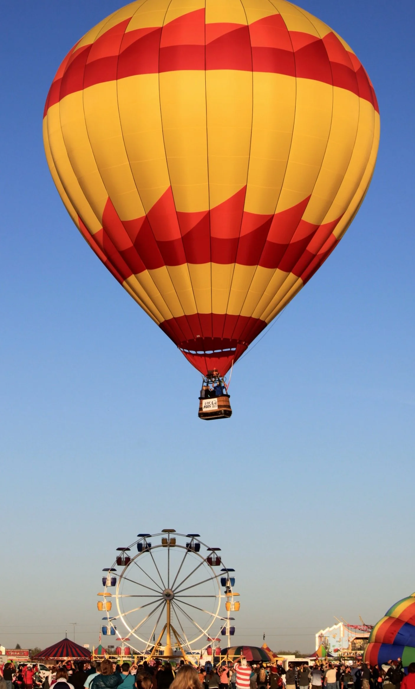 A hot air balloon with a red and yellow striped pattern flying over a carnival. The background shows a Ferris wheel and a large crowd of people walking around the fairground about to launch over the Napa Valley