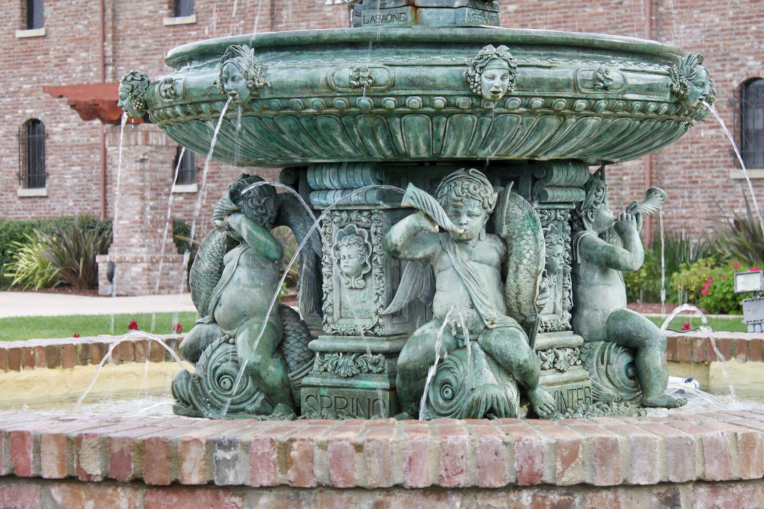 A decorative greenish bronze fountain featuring various carved faces and mythological creatures, with water flowing from some of the faces' mouths, set against a brick building background.