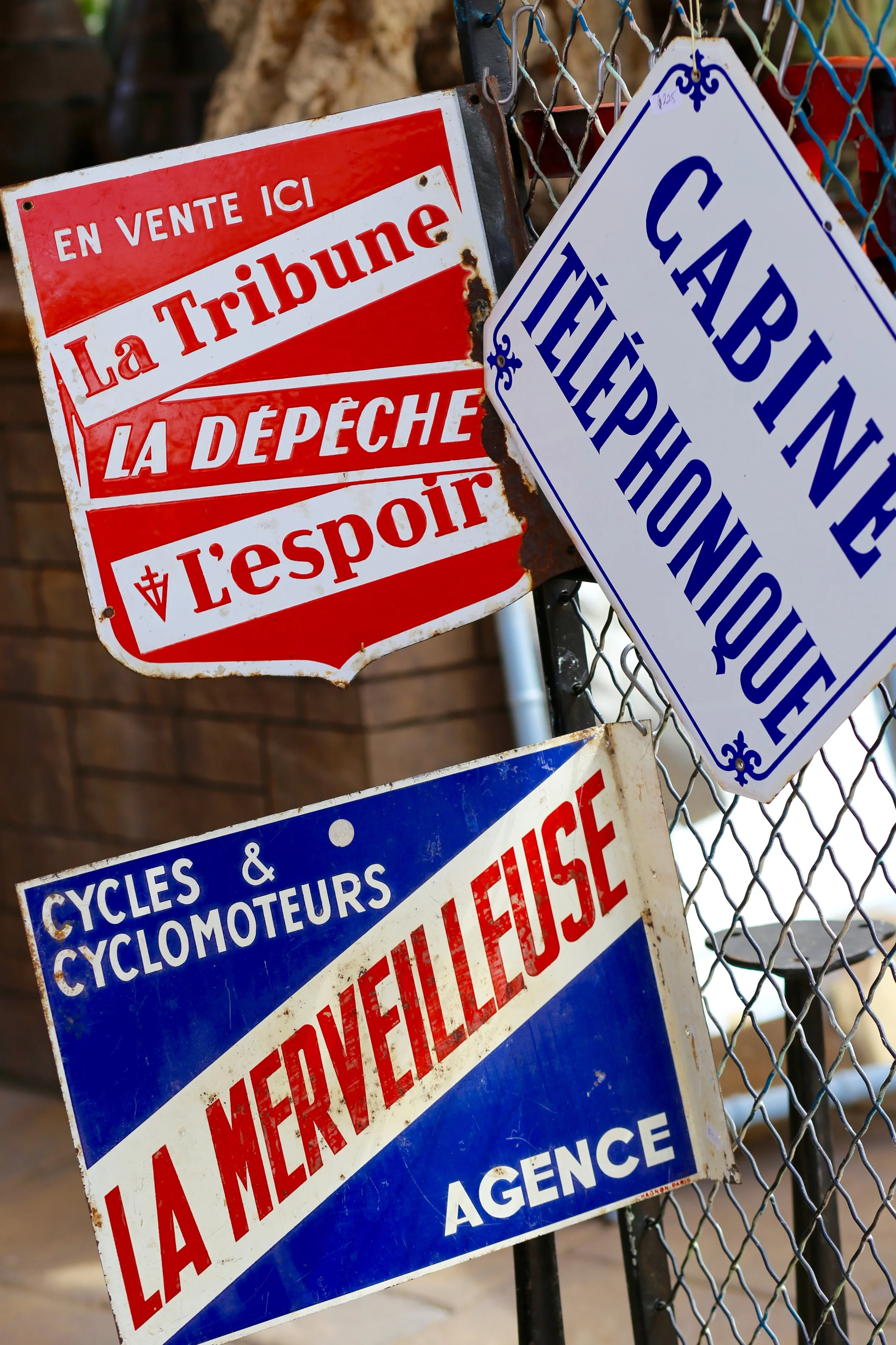 Multiple signs on a chain-link fence, including one red and white sign that reads in French, "En vente ici La Tribune La Dépêche L'Esprit," and a white and blue sign that reads "CAMPBINE" and "CYLES & CYCLAMOTEURS." The signs are slightly rusty and w