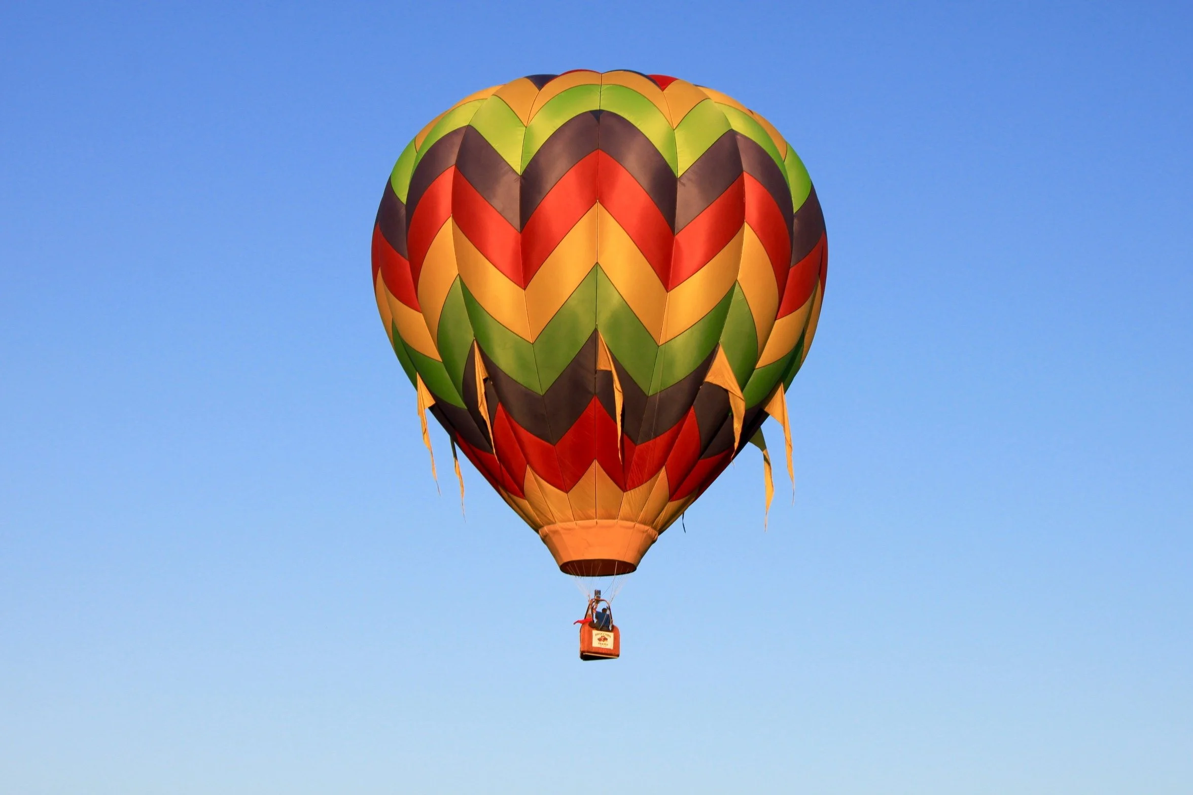 Colorful hot air balloon with a chevron pattern in orange, red, yellow, green, and black, floating in a clear blue sky.