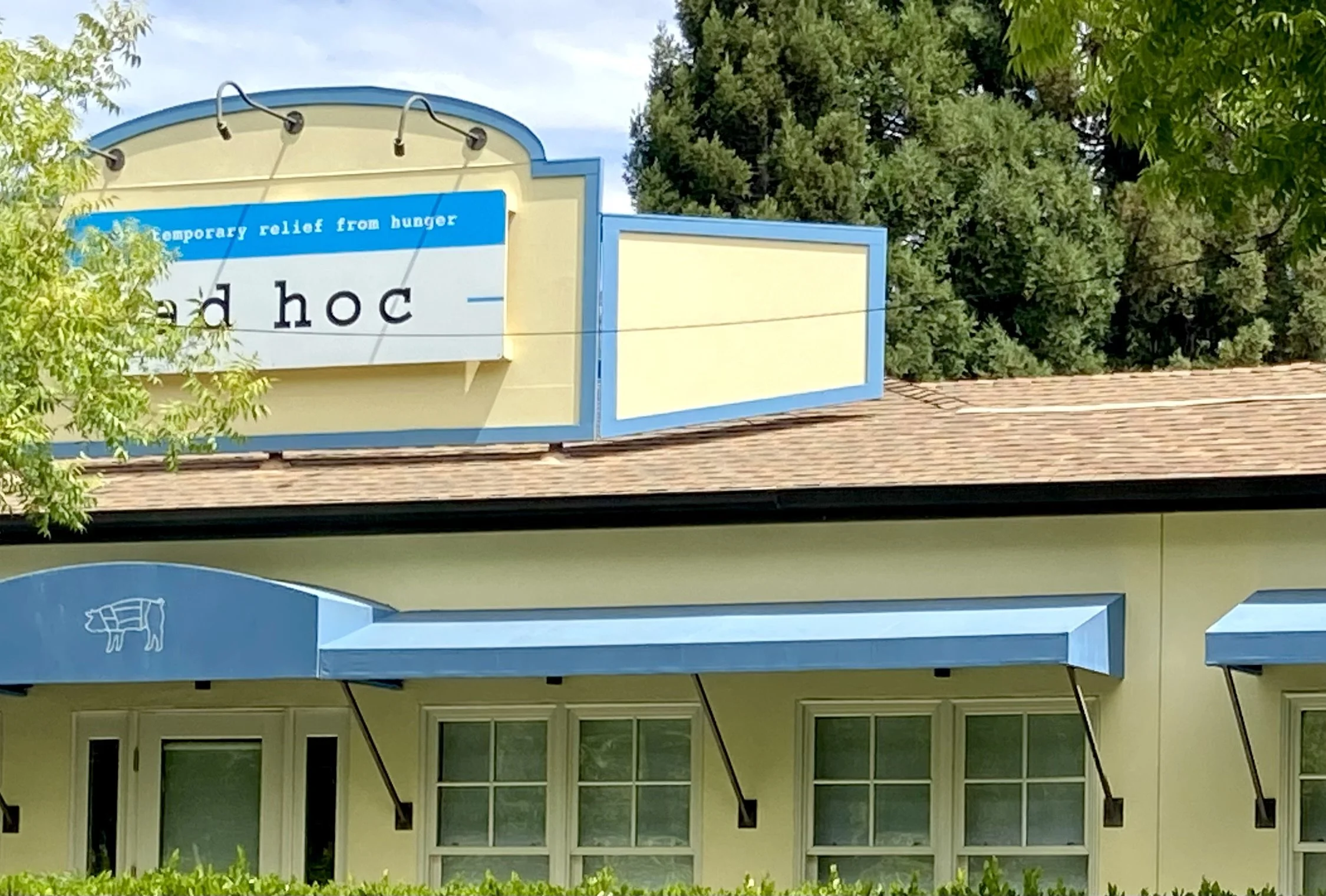 Lush green trees surround a building with a beige and light green exterior, featuring large windows and a front porch with a green hedge. Above, a blue sign of the same shape and color as the building's trim has text and a blank space for additional signage.