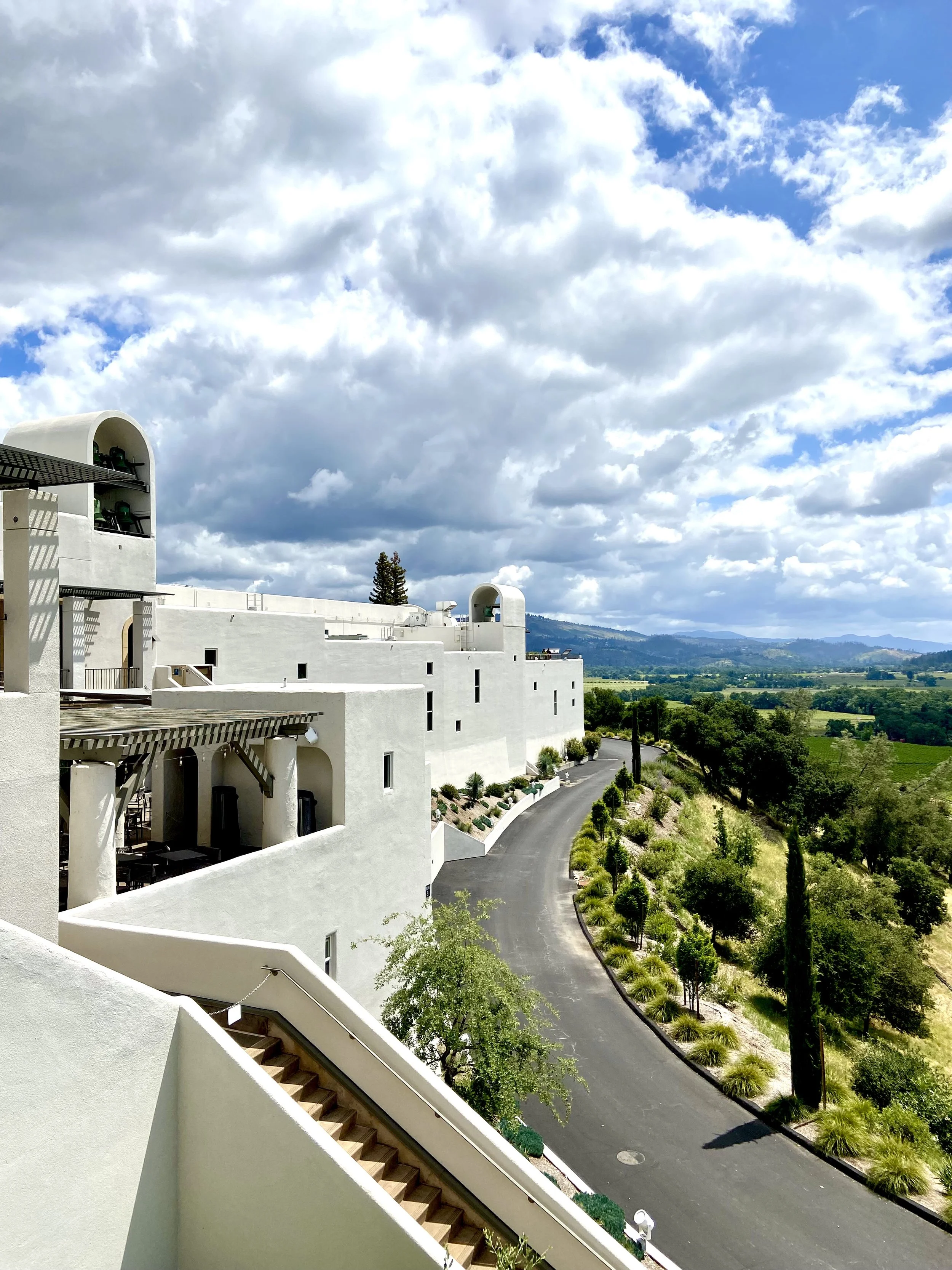 White modern building with archways and balconies, a curved road lined with greenery, trees, and landscape, under a partly cloudy sky with patches of blue.
